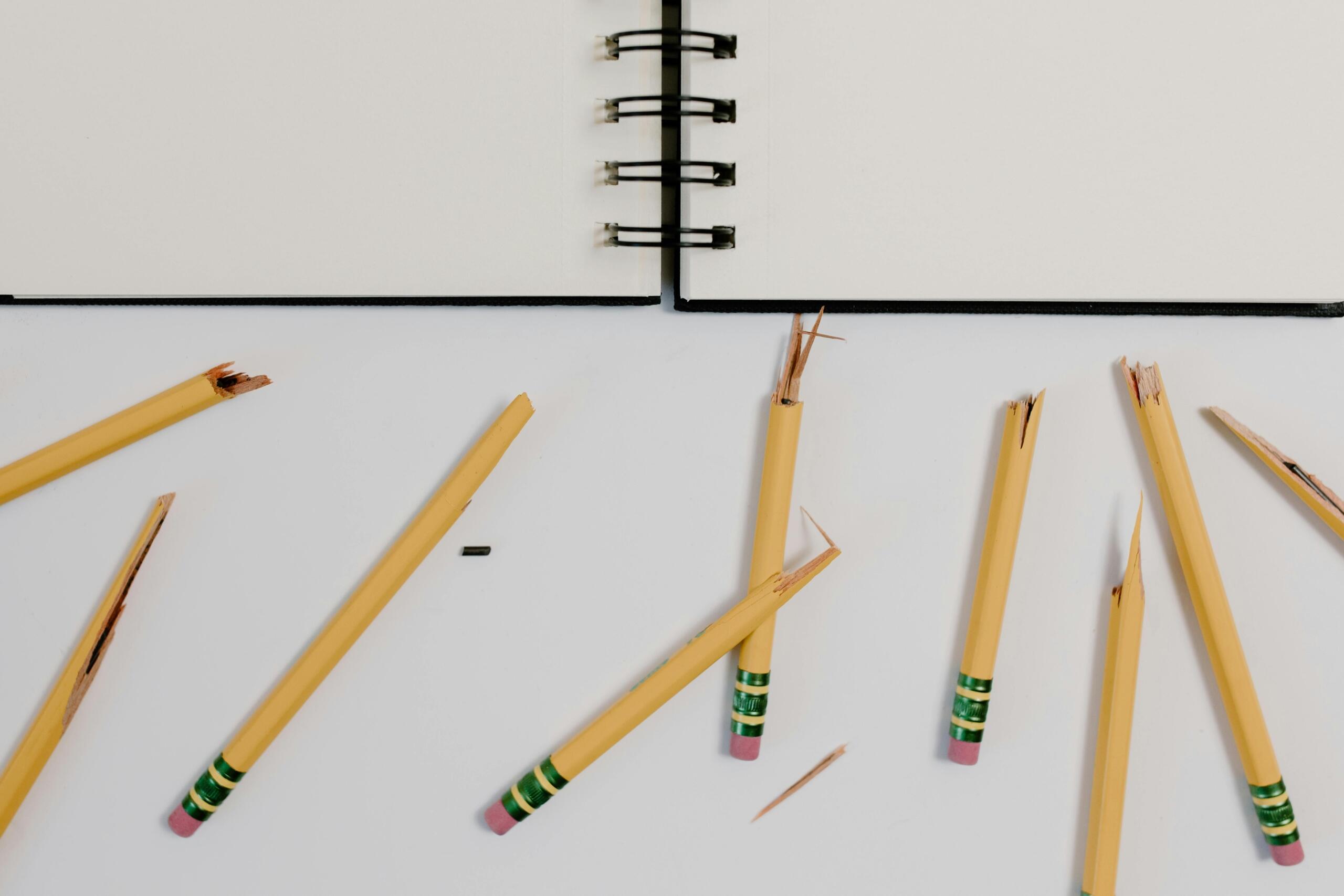 Gebroken gele potloden liggen verspreid naast een notitieboek op een wit oppervlak.