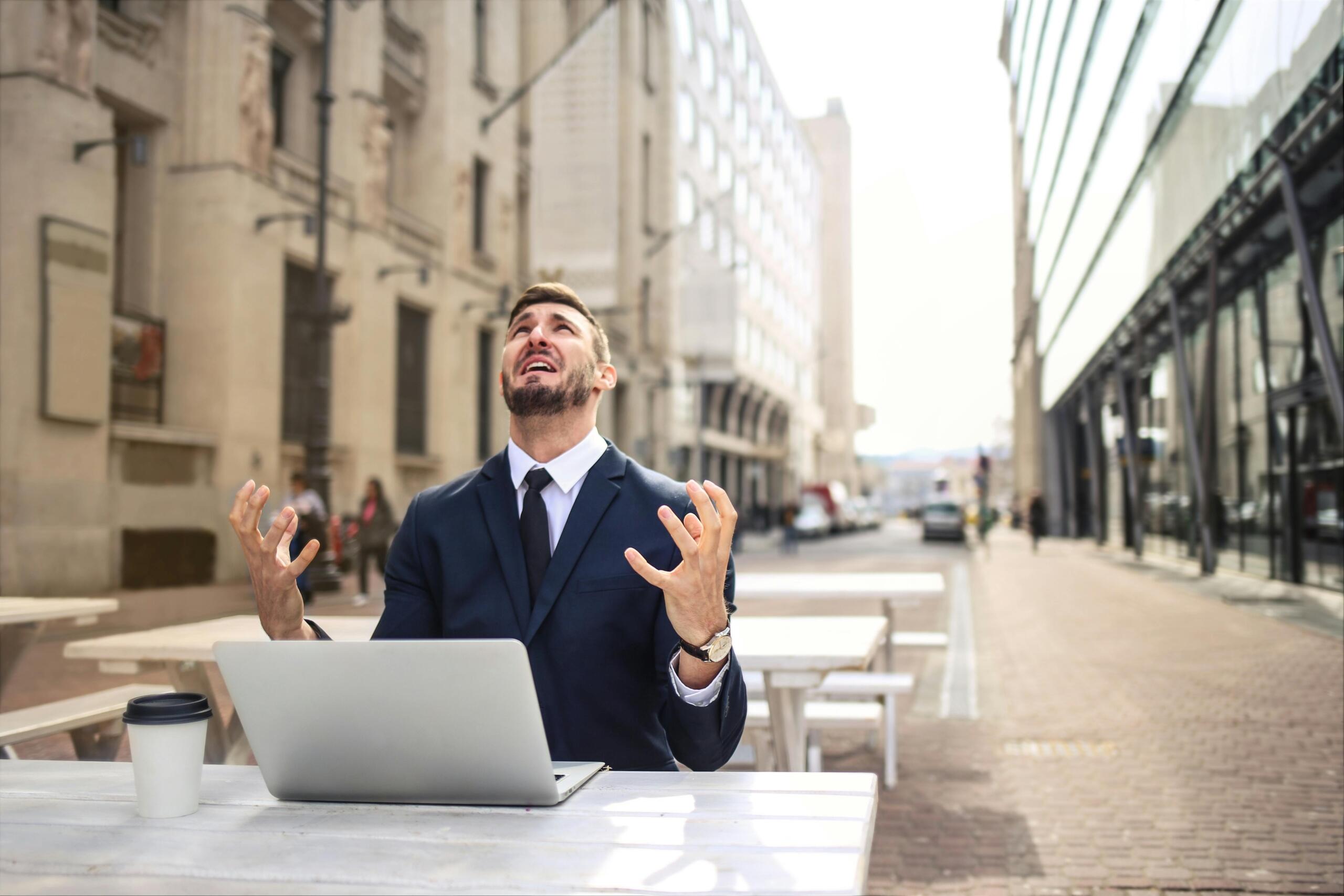 Persoon zit aan tafel buiten in een stad, achter een laptop, en heft zijn handen in paniek op.