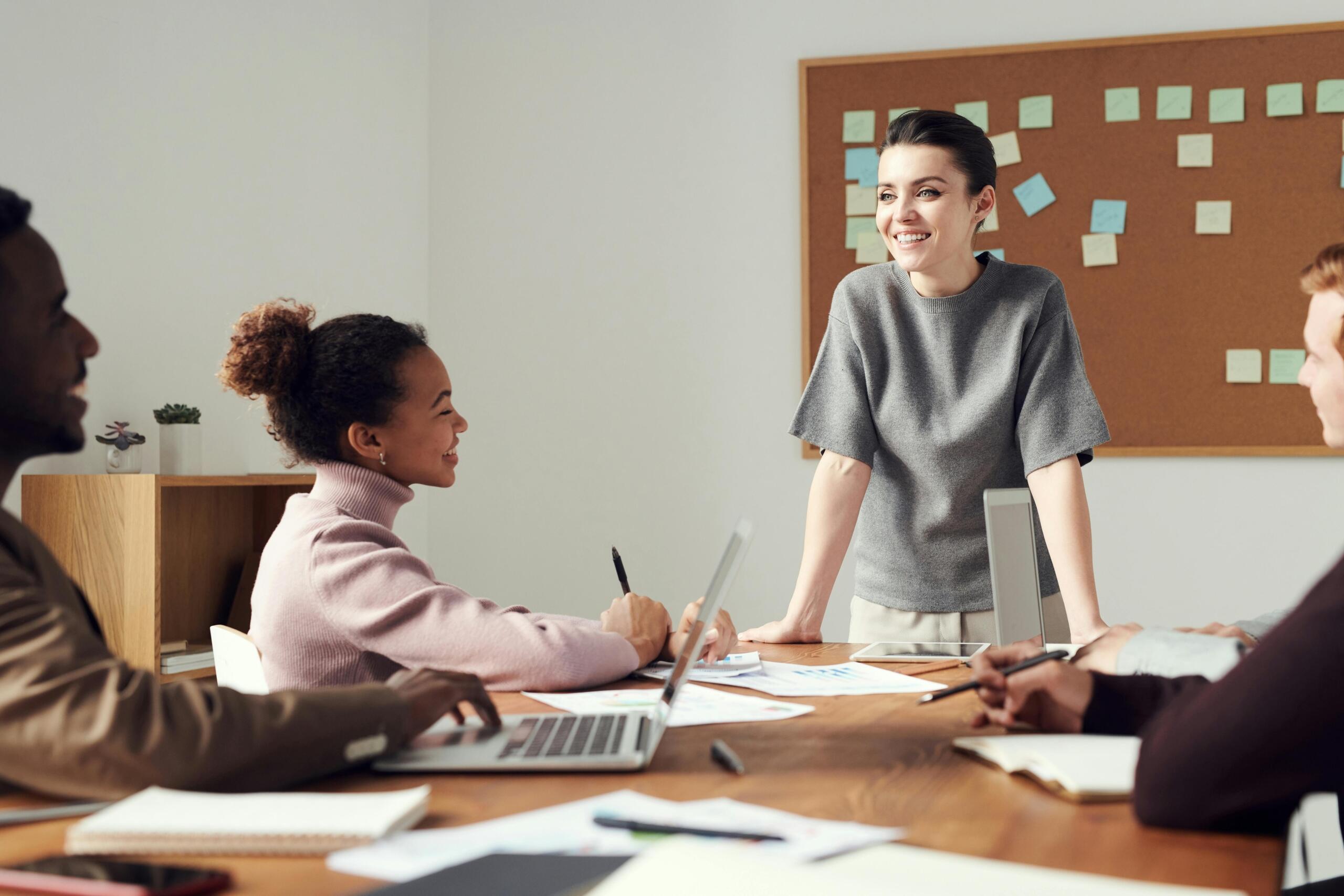 Glimlachende vrouw staat aan het hoofd van een tafel met diverse collega’s, met een prikbord vol memo’s op de achtergrond.