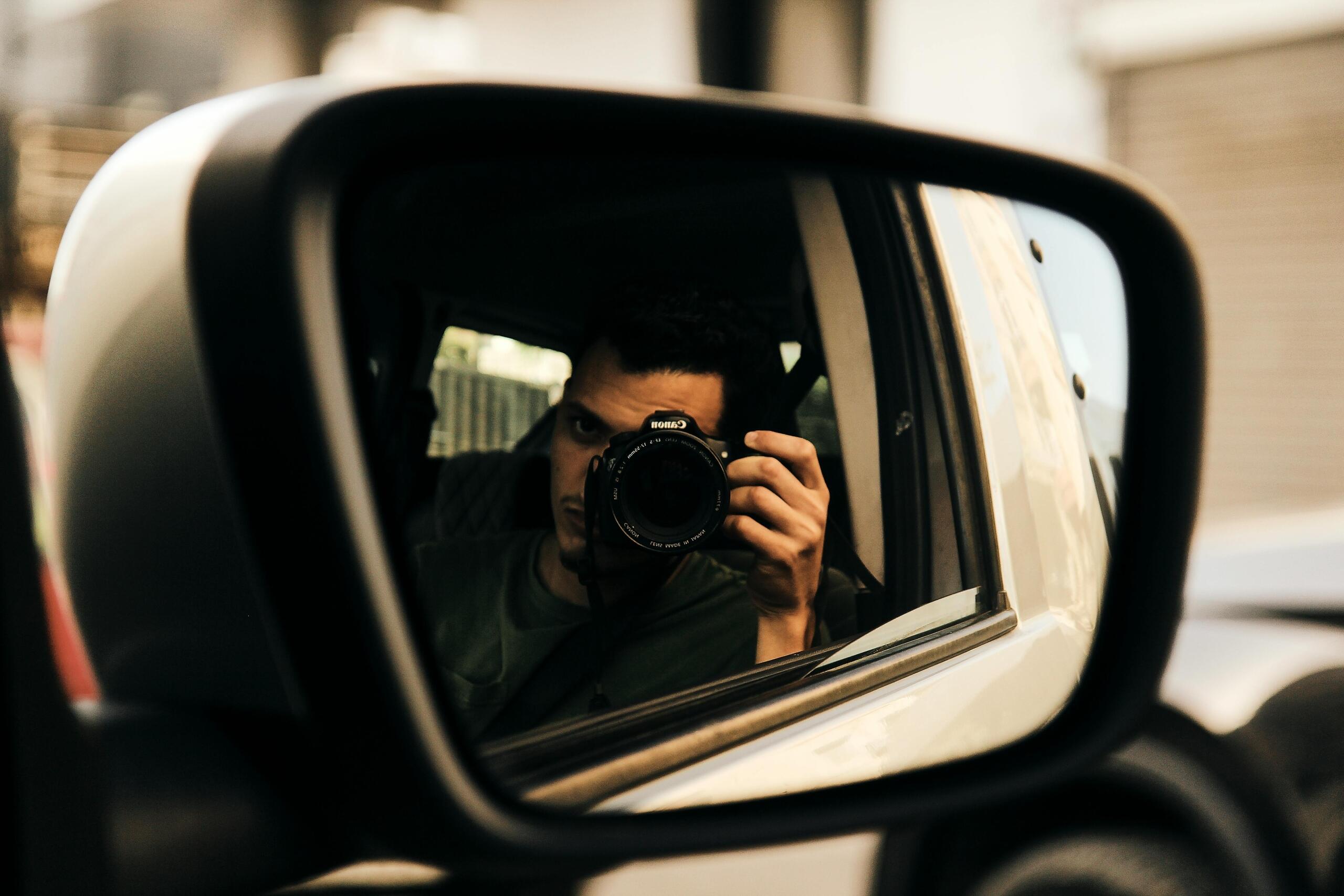 Man met camera weerspiegeld in de zijspiegel van een auto, met blurred straat op de achtergrond.