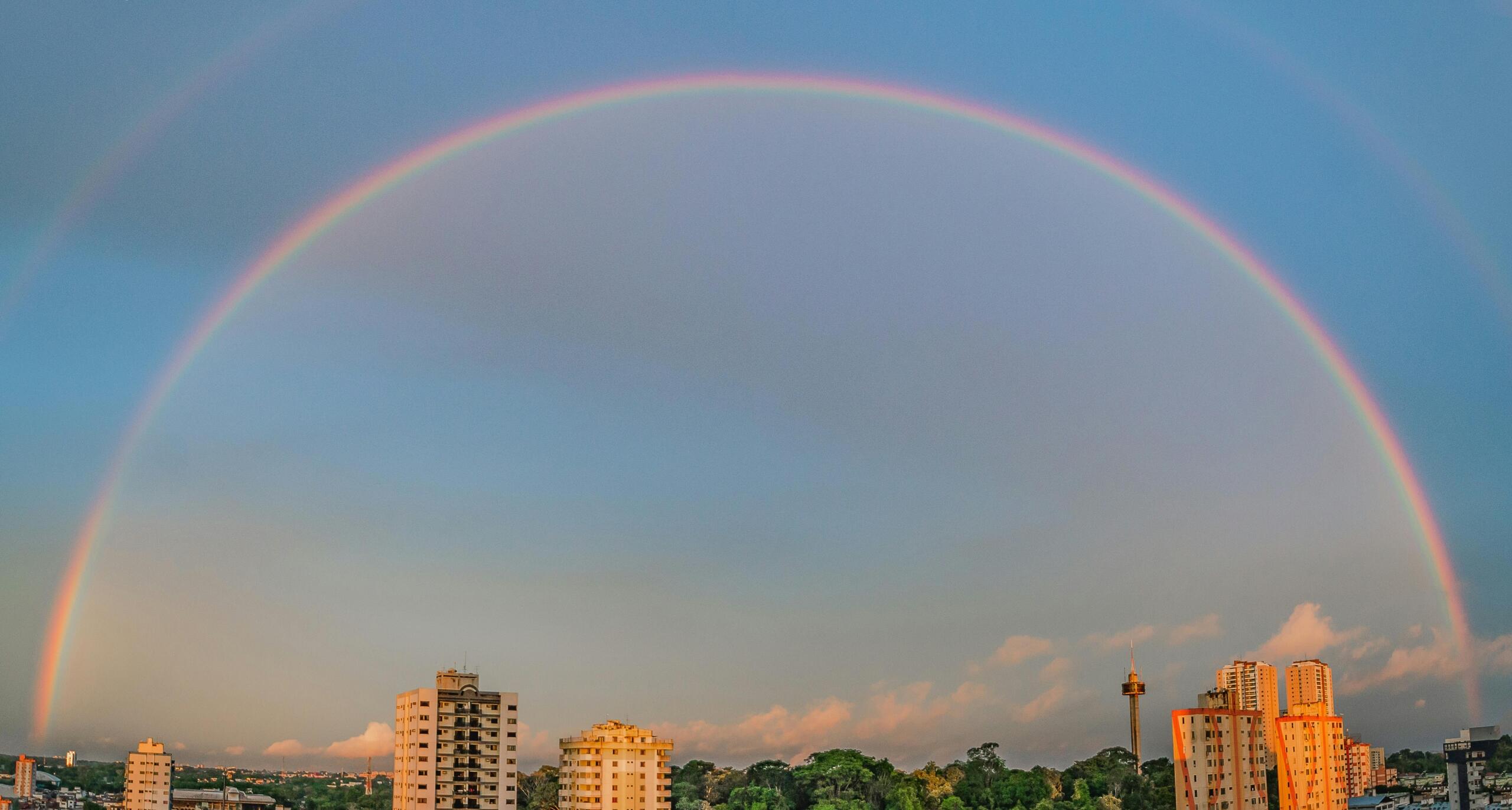 Volledige regenboogboog aan een blauwe hemel boven een stadsgezicht met hoge gebouwen en groen.