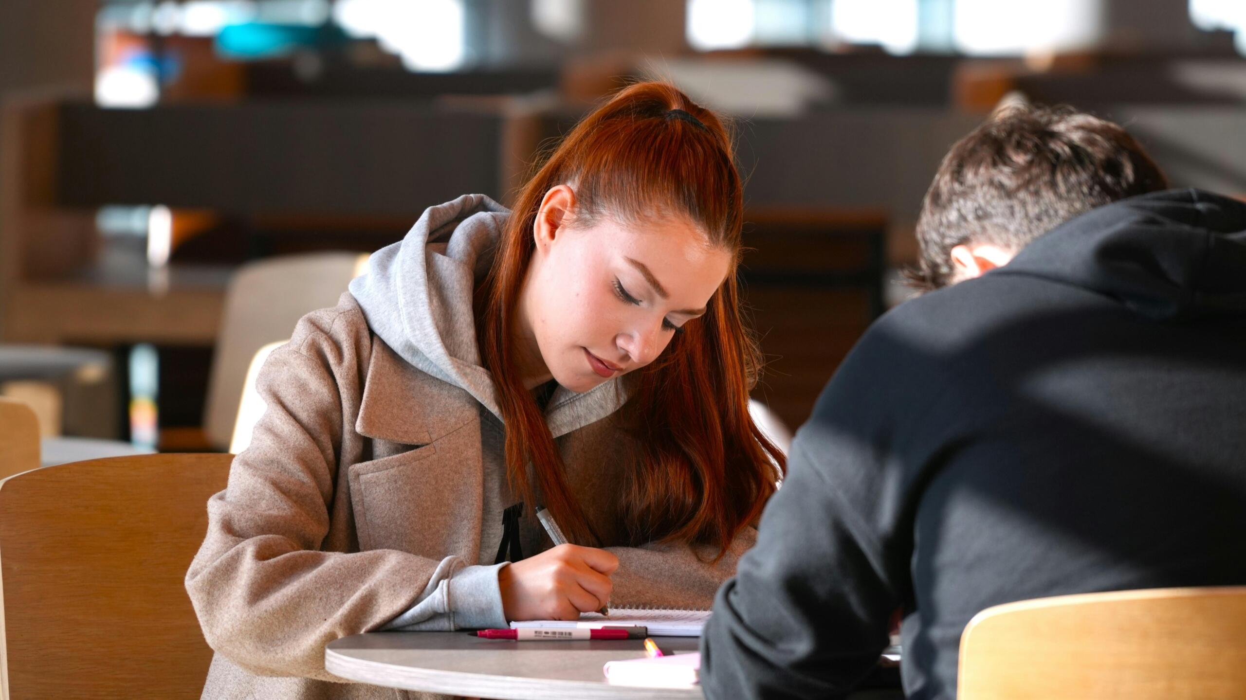 Een jonge vrouw schrijft geconcentreerd aantekeningen aan een ronde tafel terwijl een medestudent tegenover haar zit.