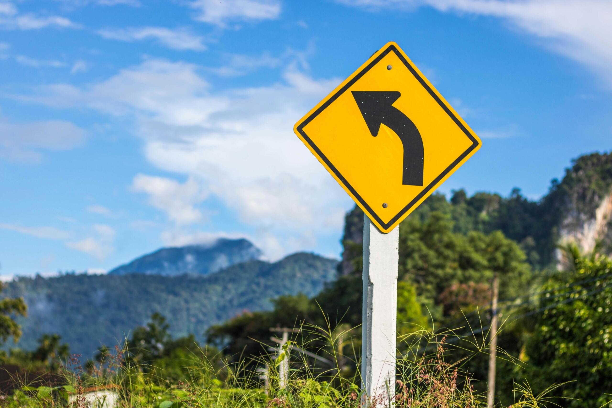 Een geel verkeersbord met een pijl naar links staat langs een plattelandsweg met bergen en groene velden op de achtergrond.