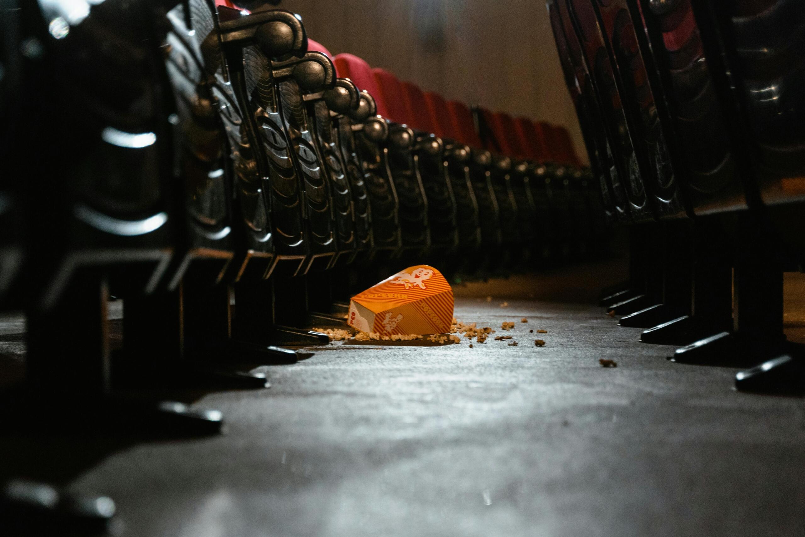 Een lege gang tussen bioscoopstoelen met een omgevallen popcornbak en verspreide popcorn op de vloer.