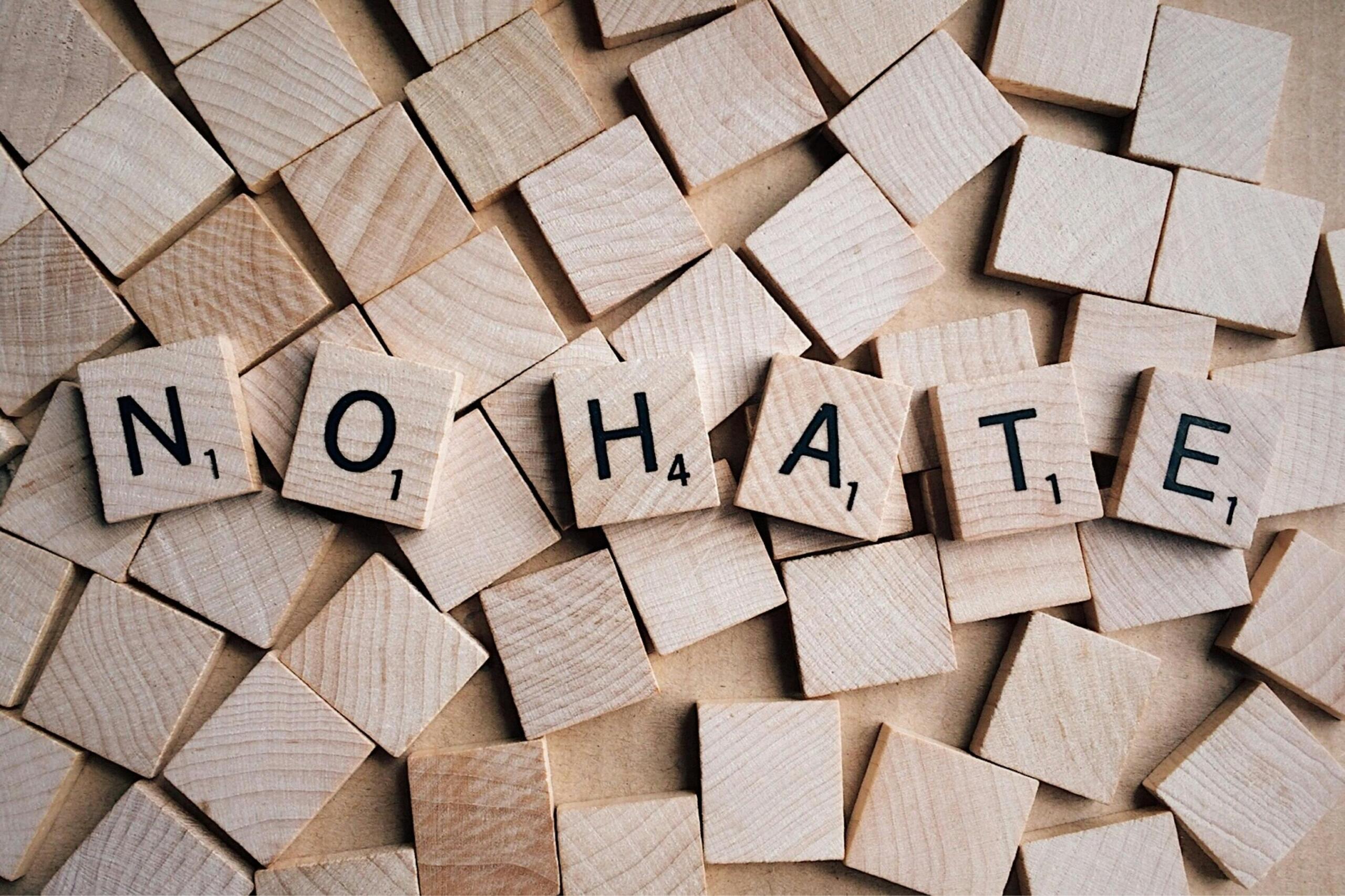 Close‑up van houten scrabble‑blokjes waarop de letters NO HATE liggen tussen stapels kleine houten blokjes.