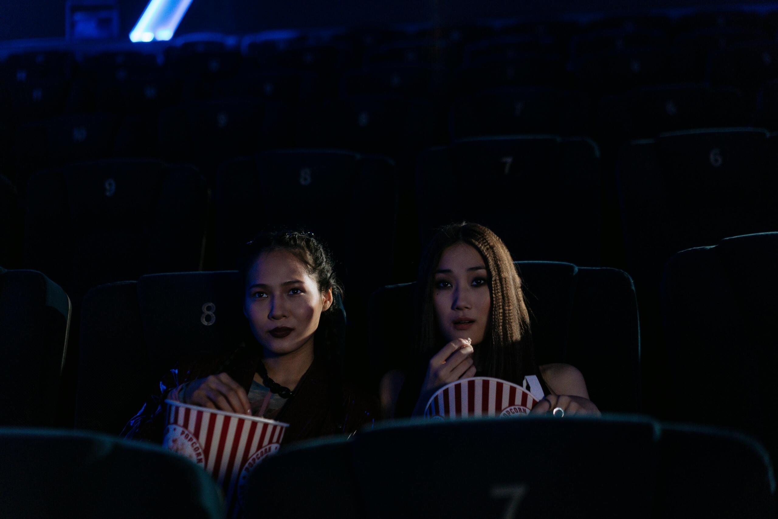 Twee jonge vrouwen zitten in een donkere bioscoopzaal met popcorn en kijken naar een film.