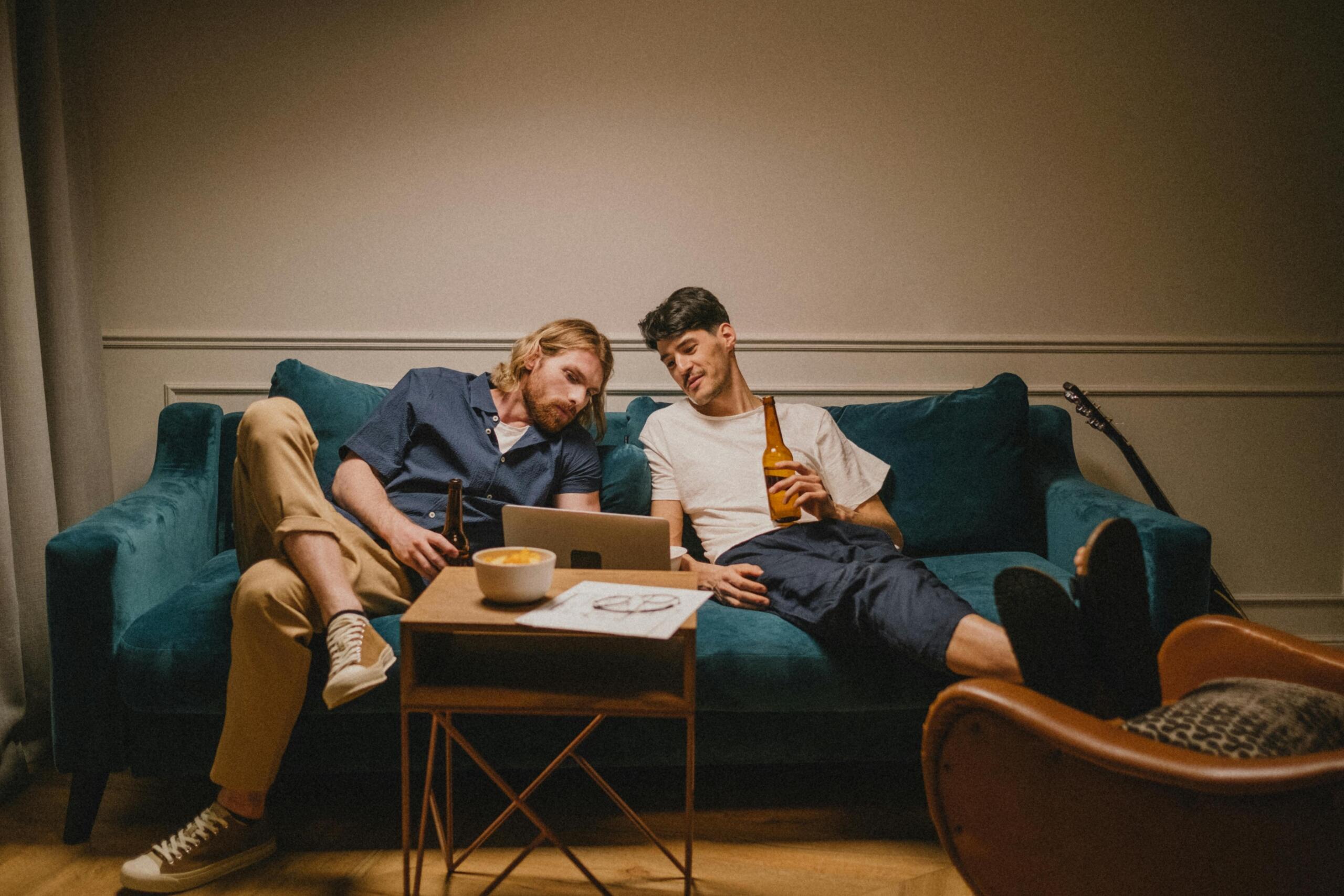 Twee vrienden hangen op een bank en kijken samen naar een laptop, met bierflesjes en snacks op tafel.