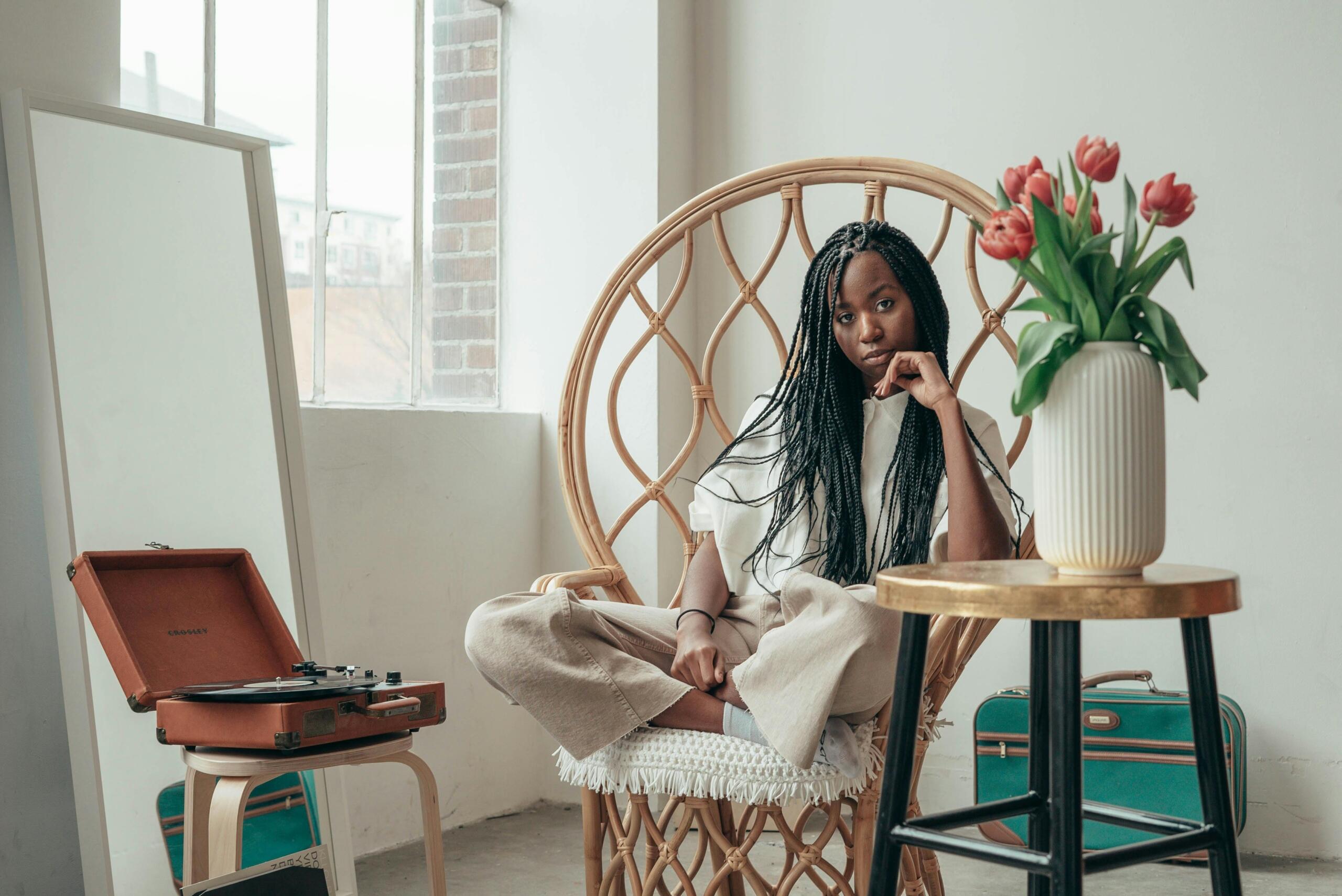 Een jonge vrouw zit in een rieten stoel naast een platenspeler en een vaas tulpen in een lichte kamer.