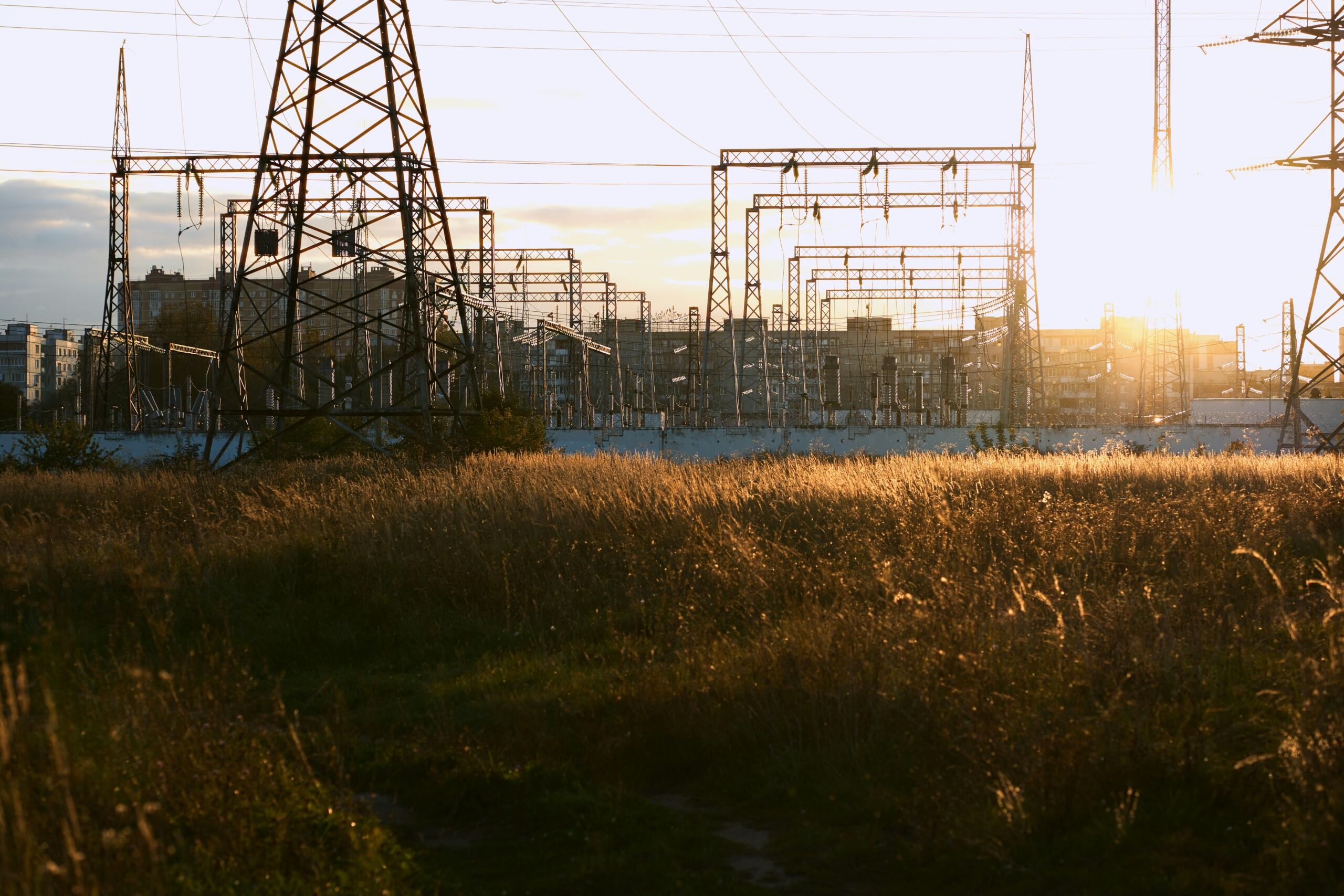 Een reeks hoogspanningsmasten en torens rijst boven een veld met droog gras terwijl de zon ondergaat.