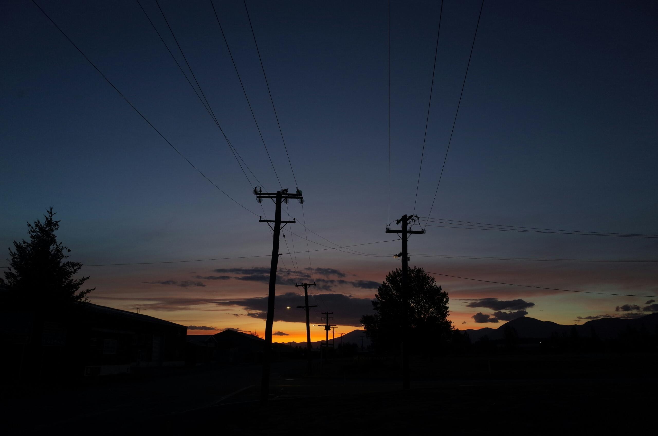 Hoogspanningslijnen en palen silhouetteren tegen een kleurrijke zonsondergang, met bergen op de achtergrond.