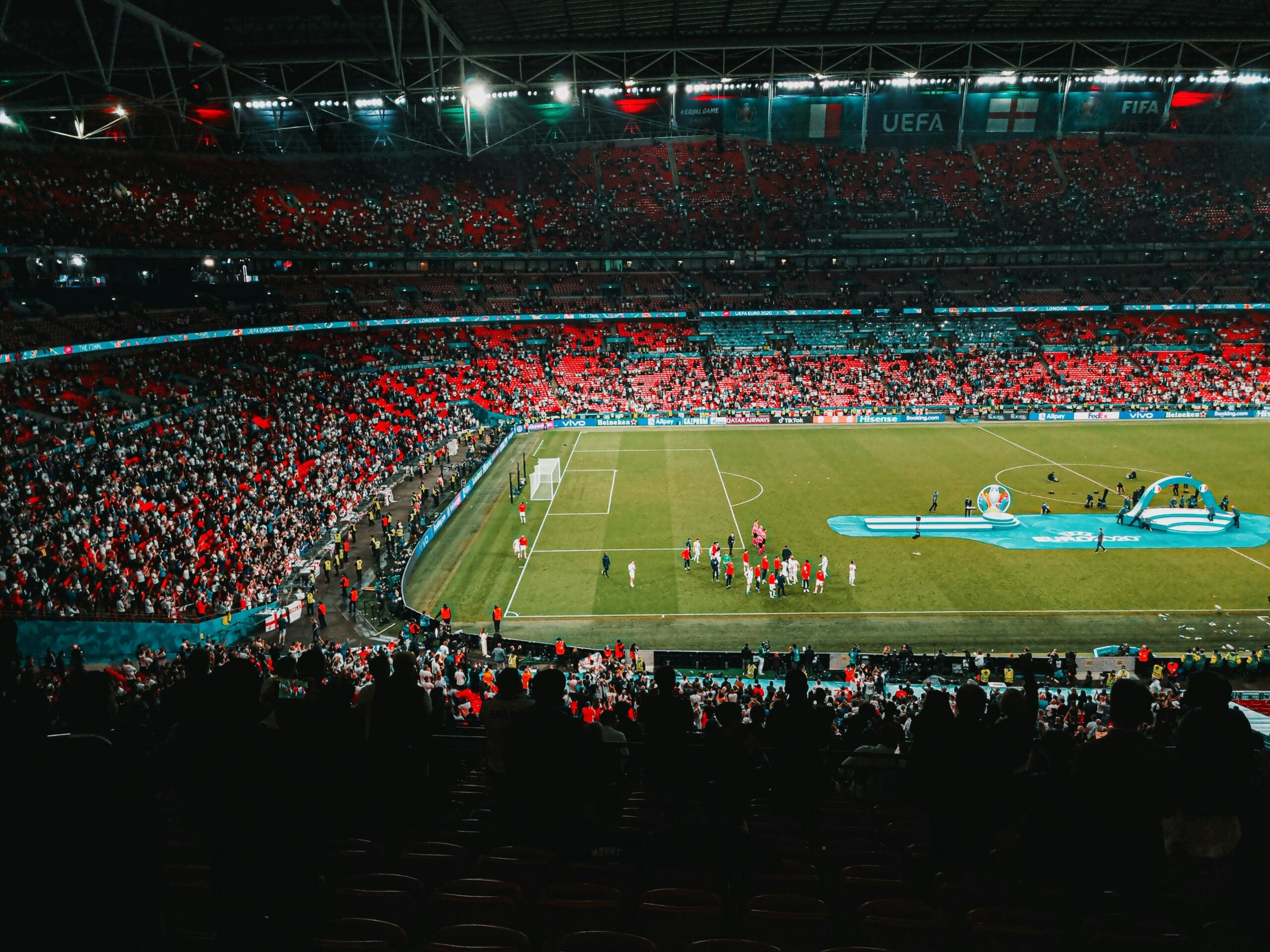 Een verlicht voetbalstadion met supporters op de tribunes tijdens een wedstrijd.