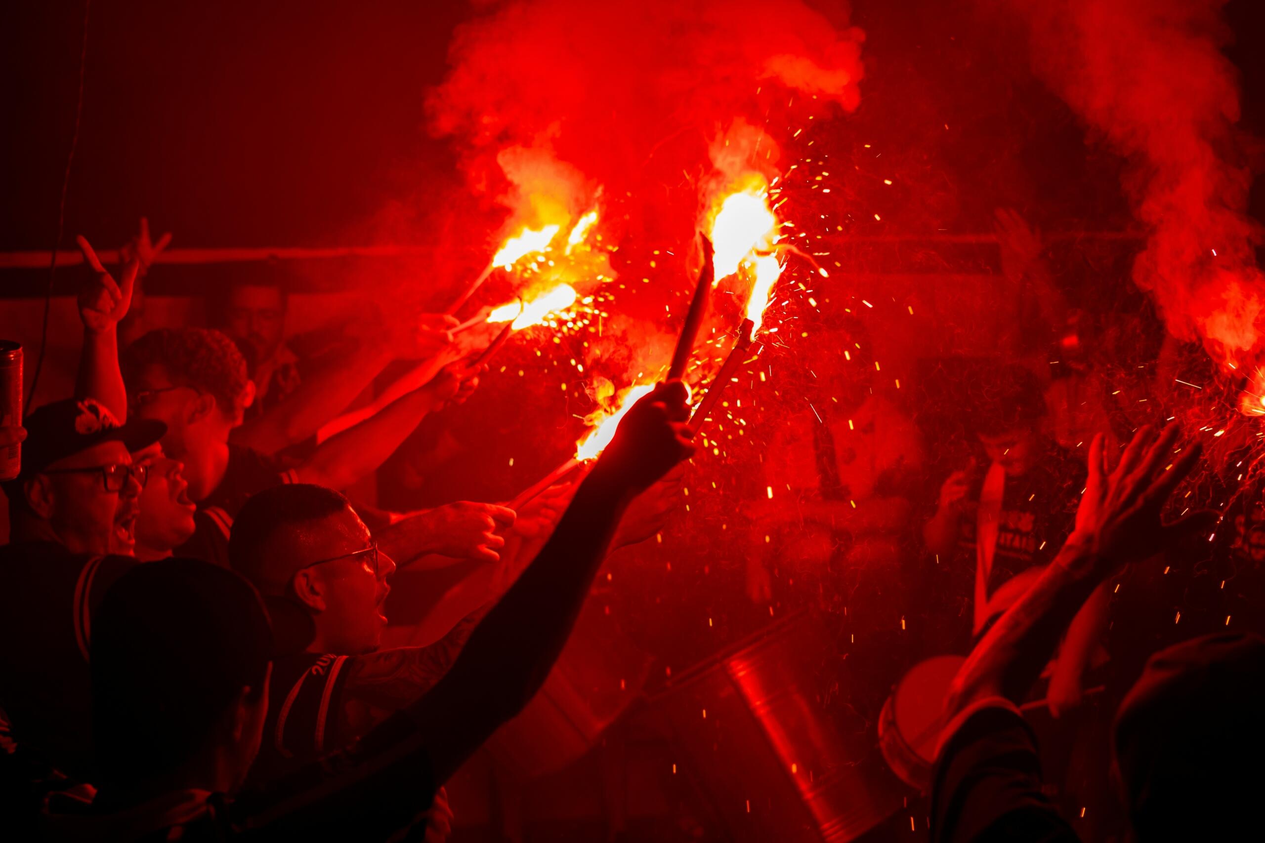 Supporters houden rode fakkels omhoog in een stadionachtige omgeving in het donker.