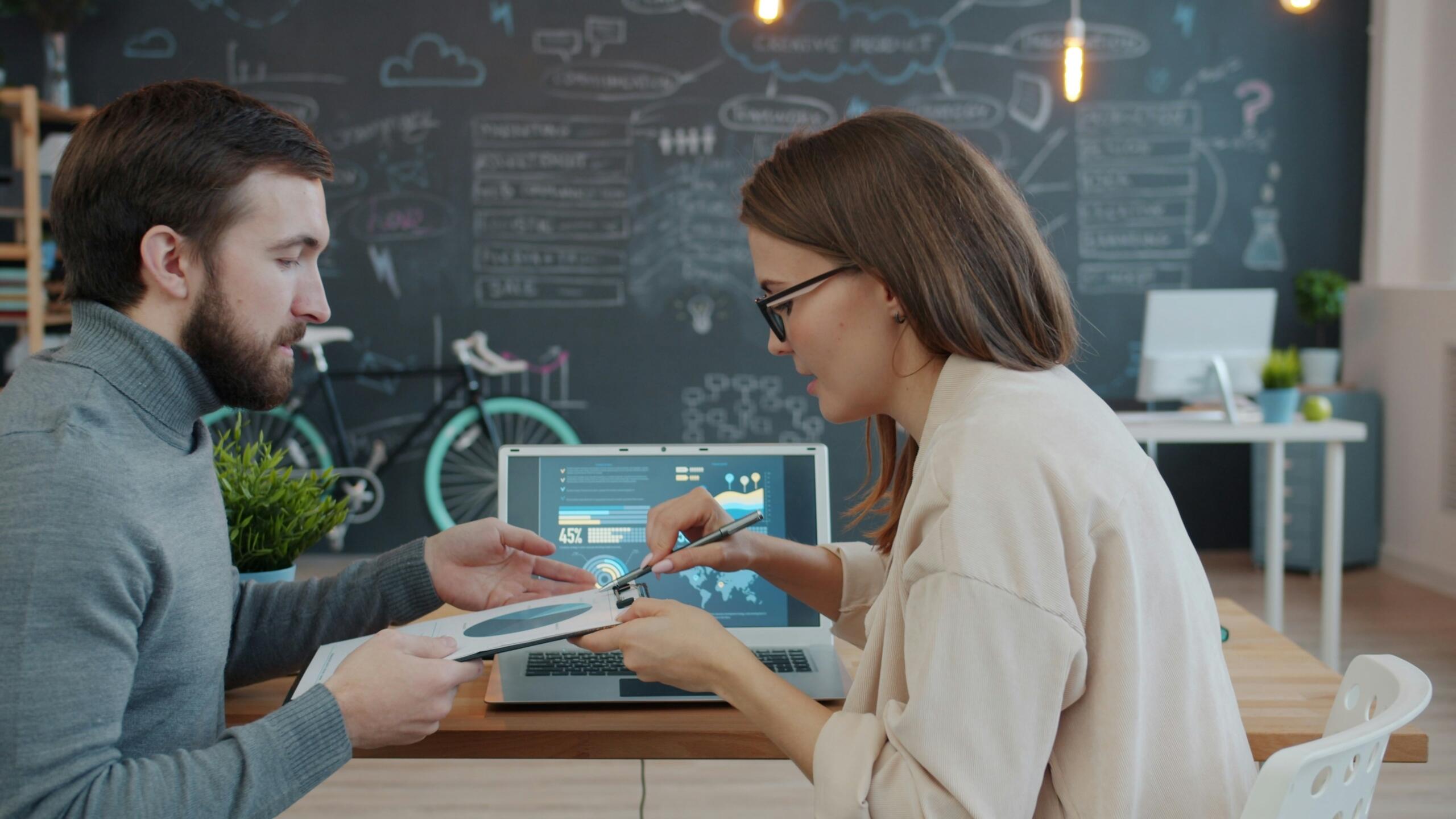 Twee collega’s zitten aan een bureau en bespreken informatie op een laptop en papier.