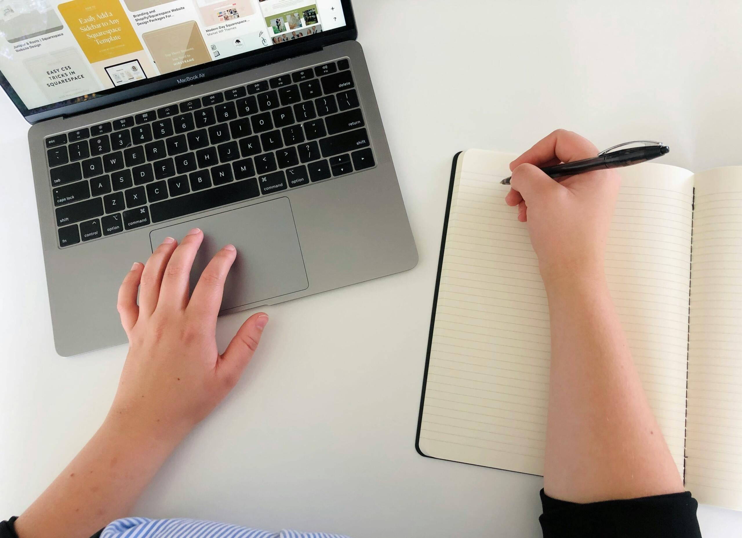 Een persoon typt op een laptop en schrijft tegelijk in een notitieboek.