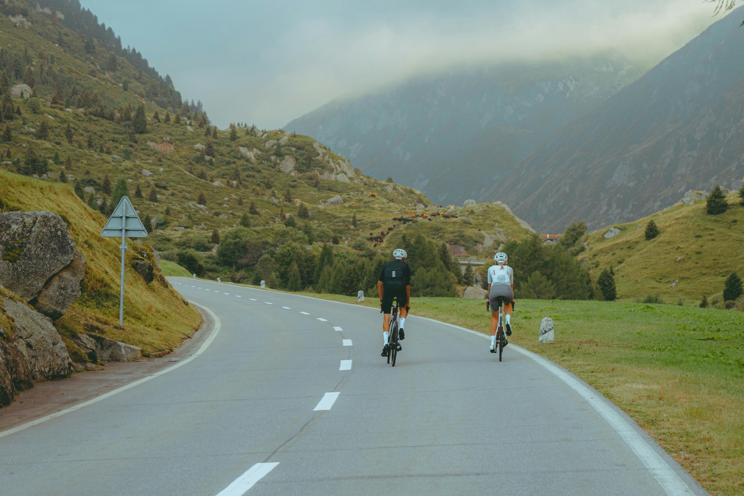 Twee wielrenners rijden over een bergweg met bergen op de achtergrond.