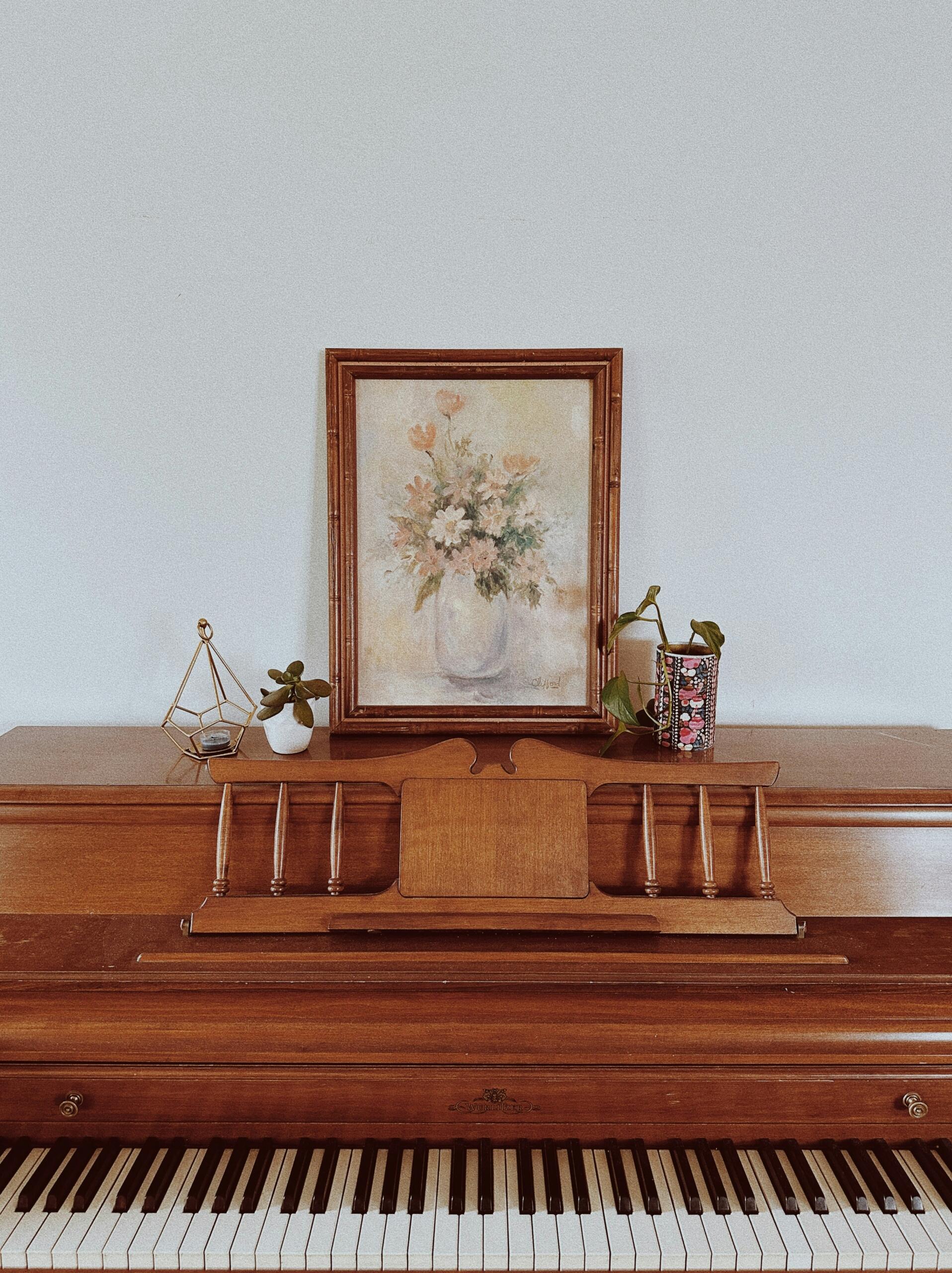 Vierkante afbeelding van een vintage upright piano tegen een lichte muur, met een ingelijste bloemenafbeelding erboven.