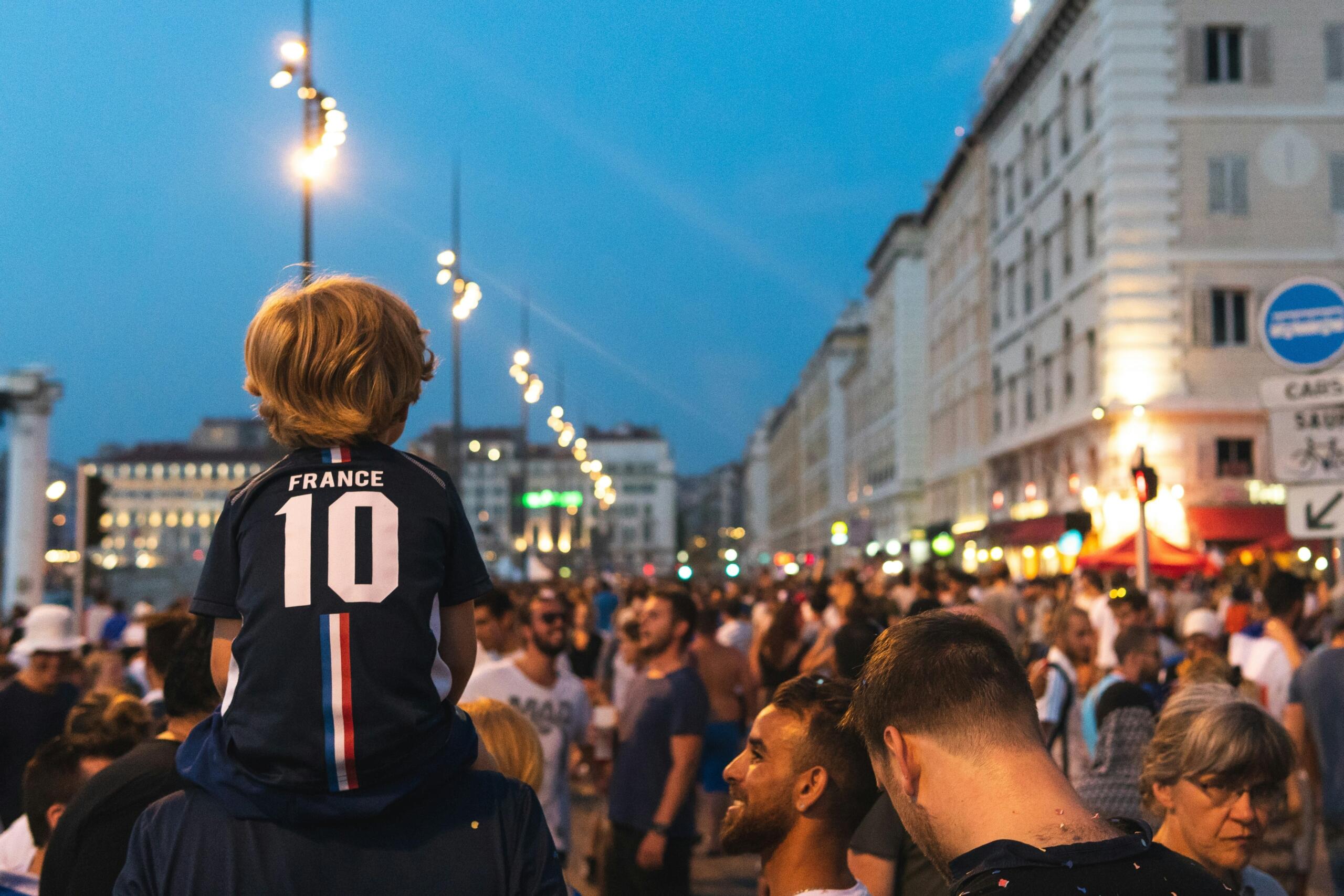 Kind met een Frans voetbalshirt op de schouders van een volwassene tussen een menigte supporters | Bron: Matthieu Joannon, via Unsplash