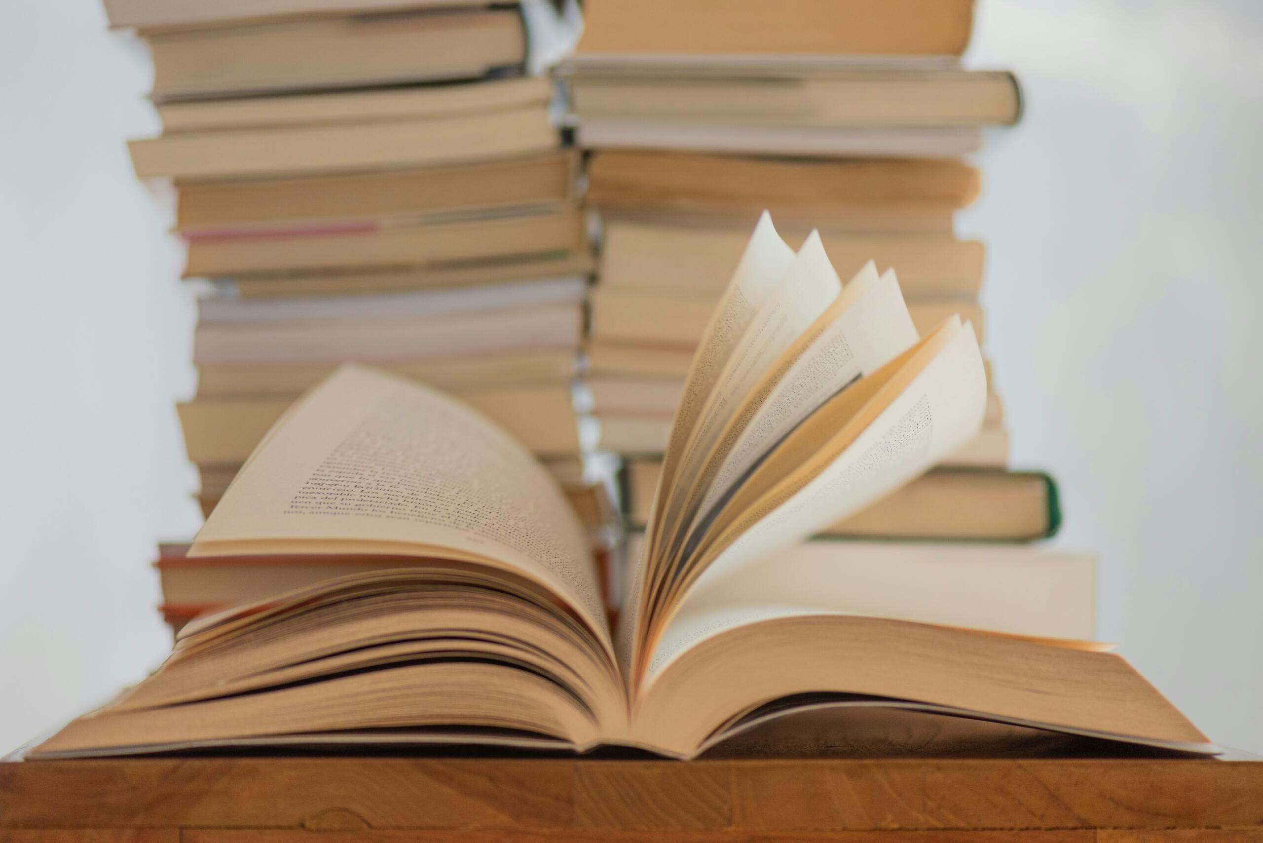 Open boek op een houten tafel met verschillende stapels boeken op de achtergrond | Bron: Elisa Calvet B., via Unsplash