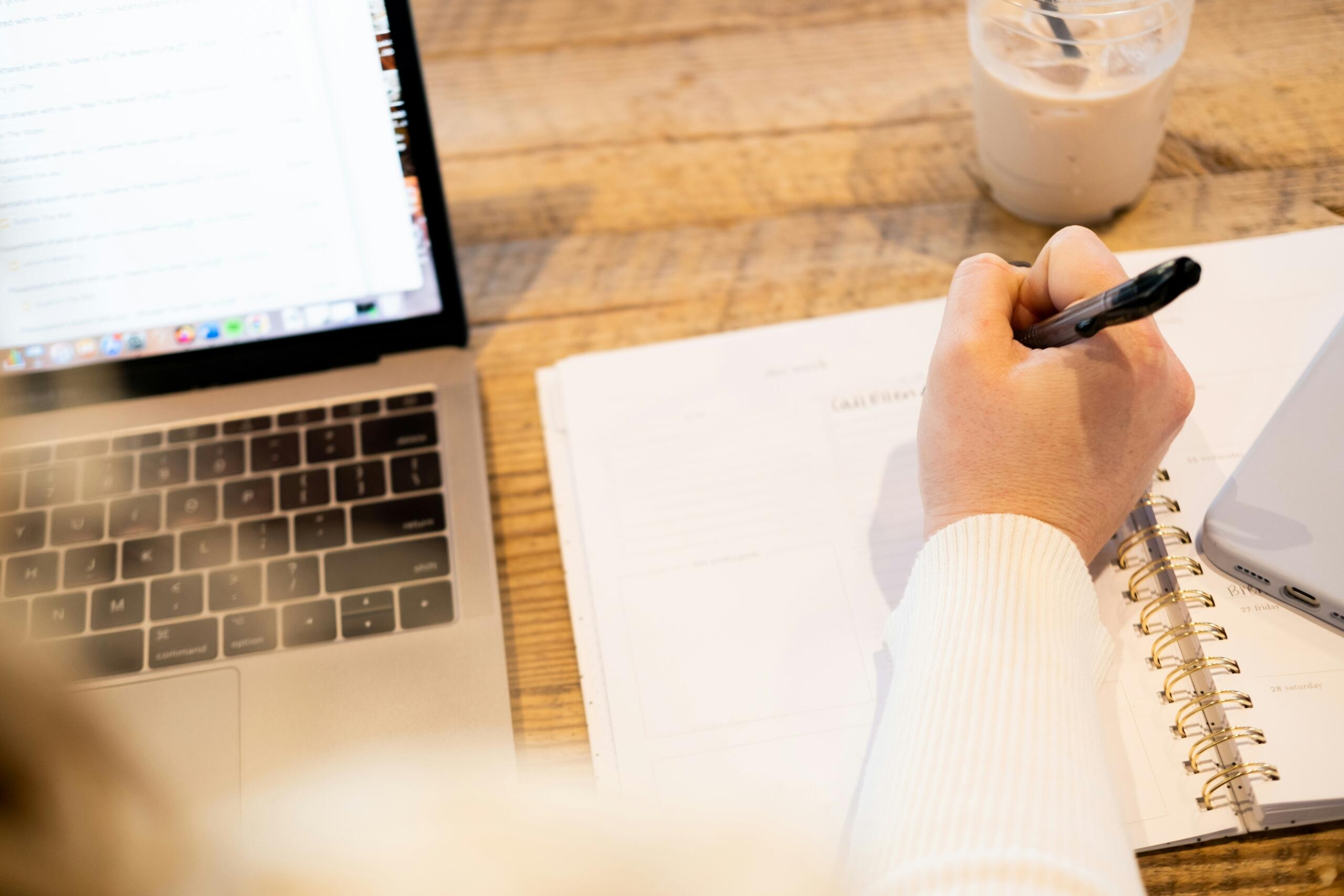 Een hand schrijft met een pen in een notitieboek naast een laptop en een glas.