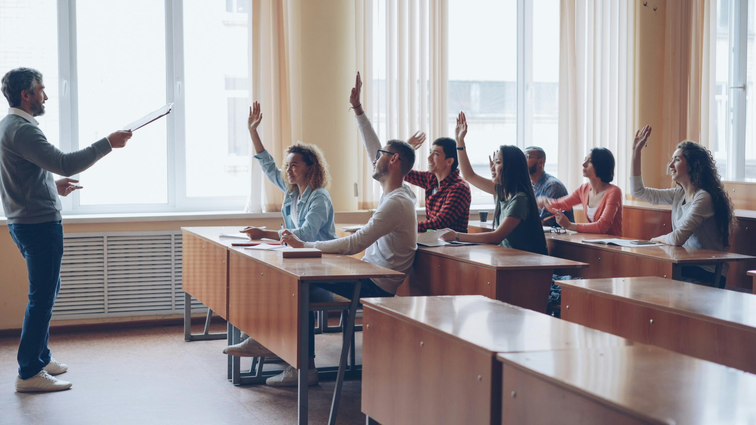 Een docent staat voor een klas terwijl meerdere studenten aan tafels hun hand opsteken.