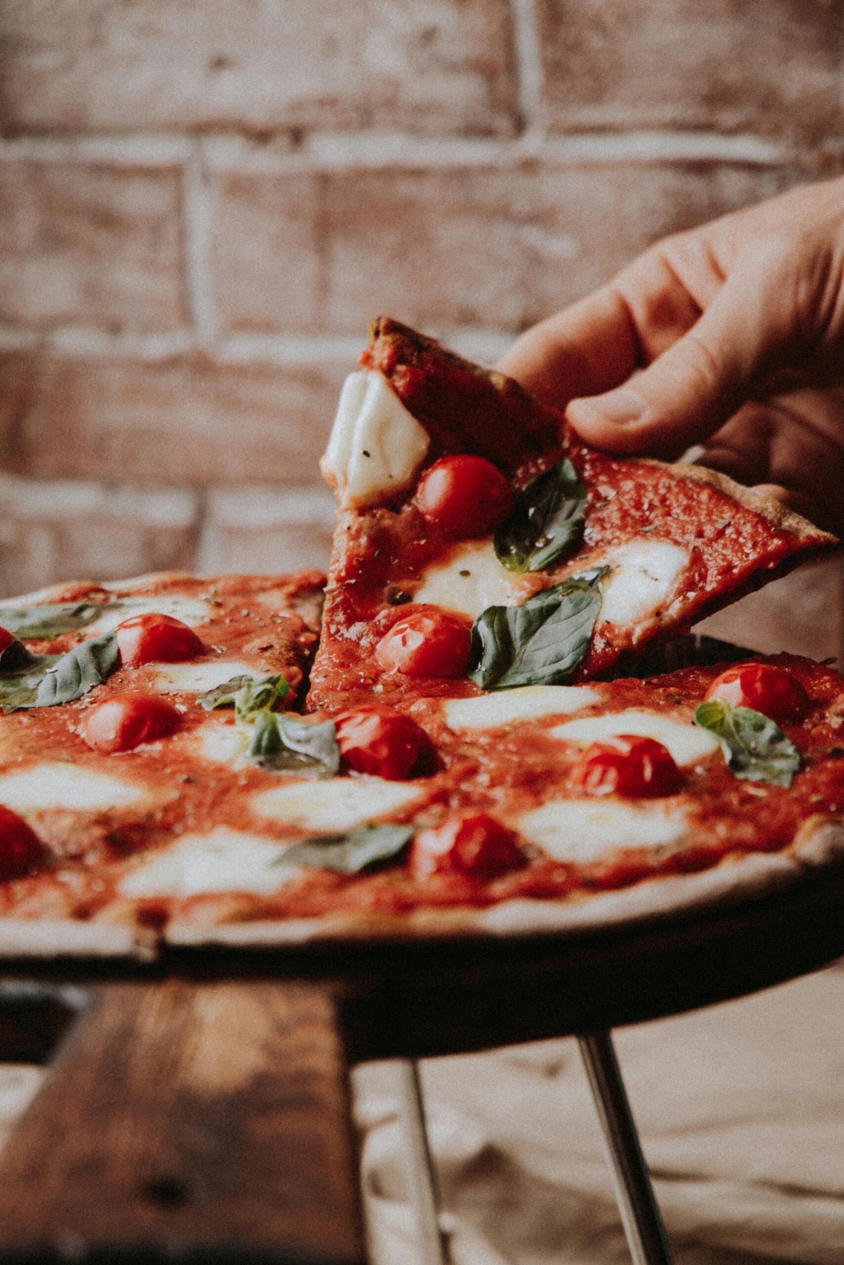 Hand pakt een punt verse pizza met tomaat, mozzarella en basilicum | Bron: Klara Kulikova, via Unsplash