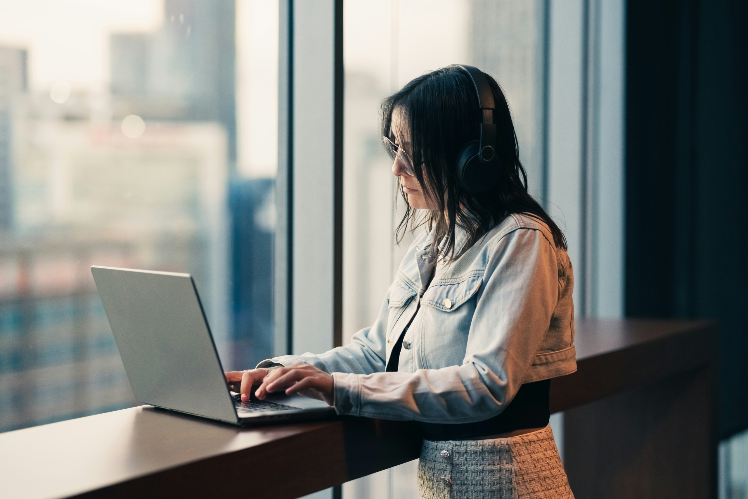Een vrouw met een koptelefoon werkt op een laptop bij een groot raam.