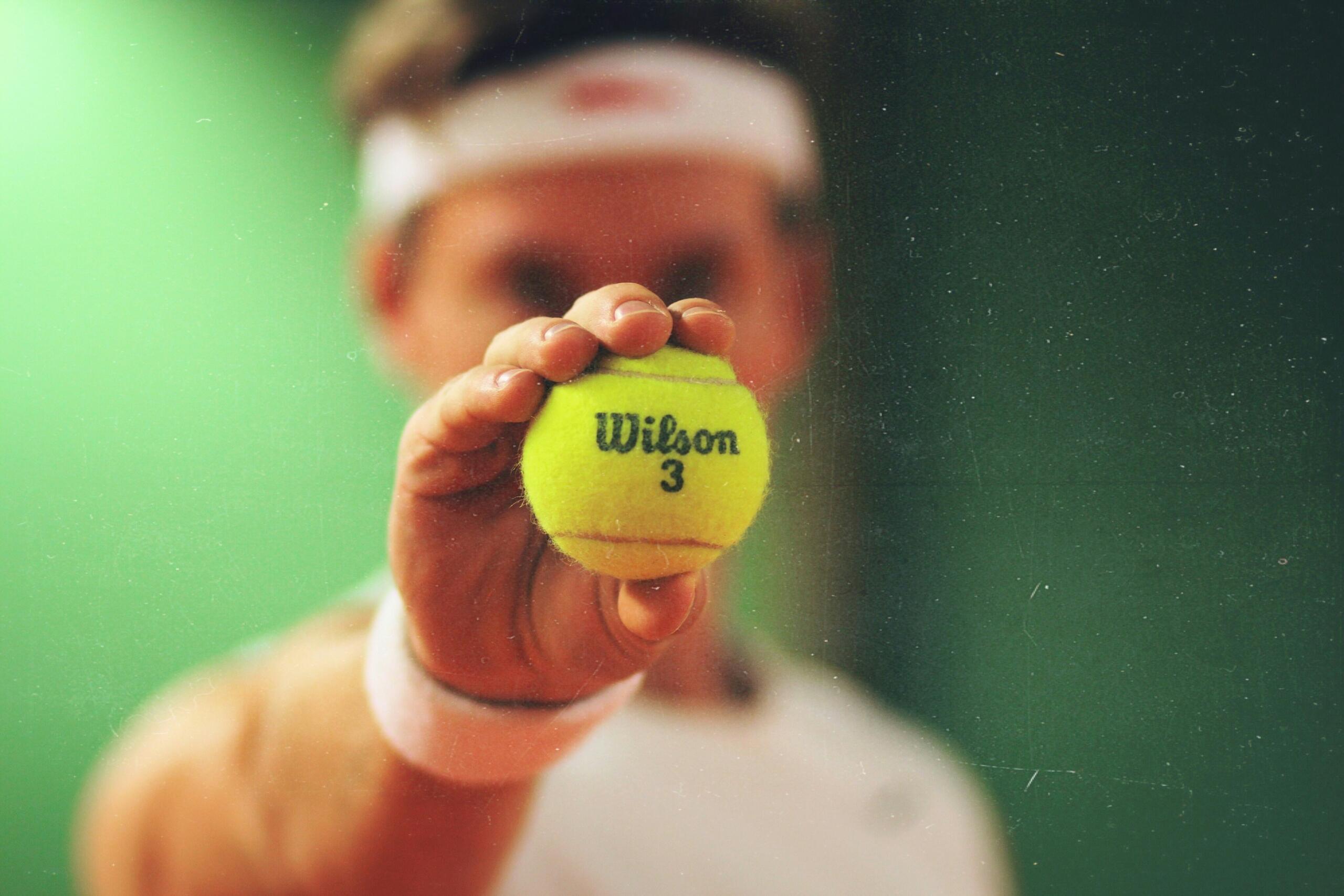 Een hand houdt een gele tennisbal dicht bij de camera, met een onscherpe tennisser op de achtergrond.