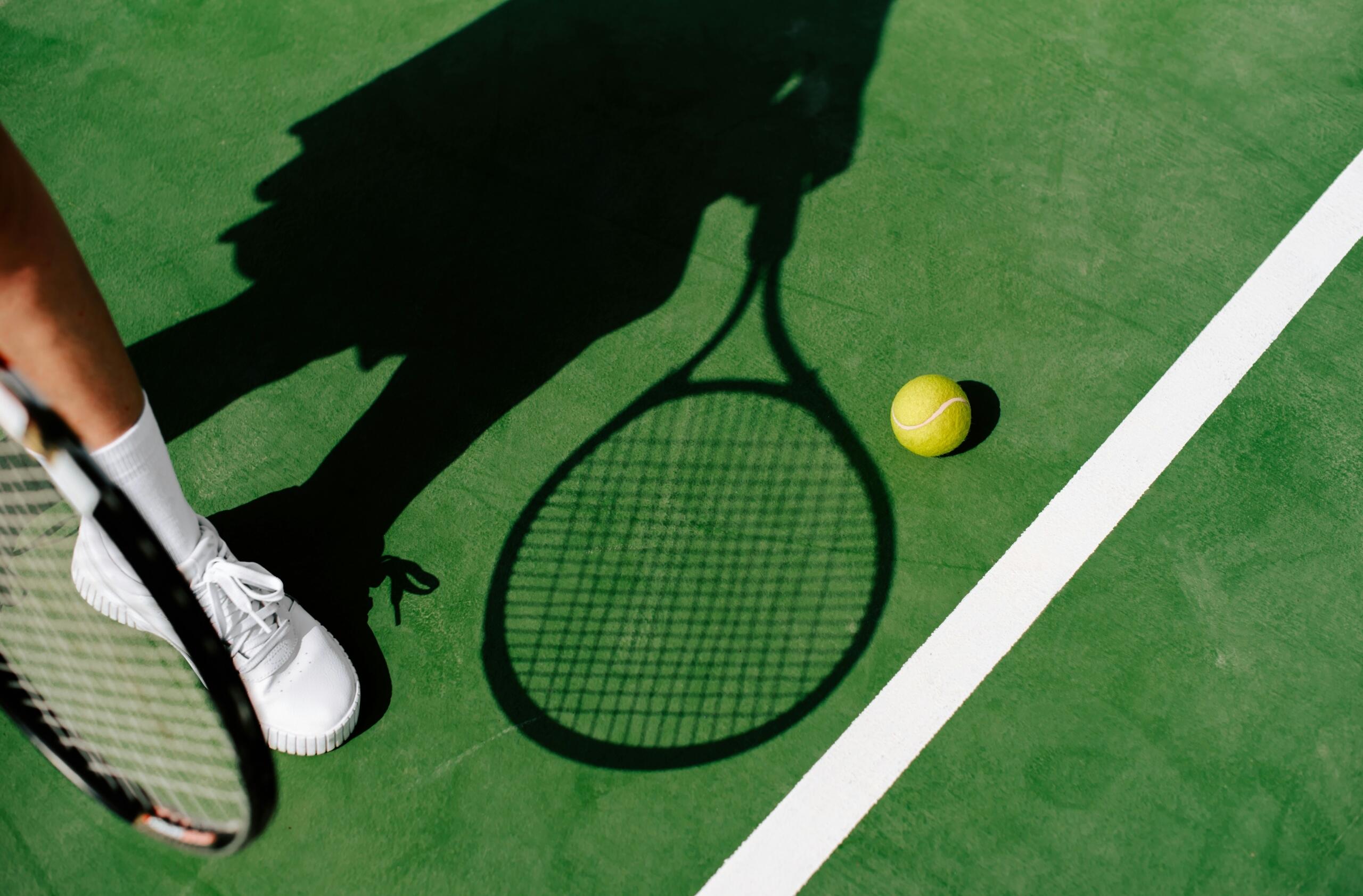 De schaduw van een tennisser met racket valt op een groene tennisbaan naast een tennisbal.