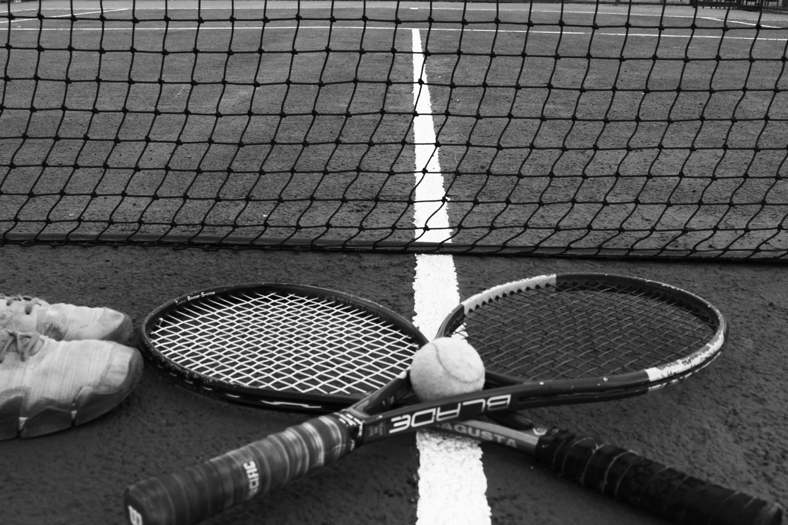Twee tennisrackets en een tennisbal liggen op een baan naast een paar tennisschoenen.