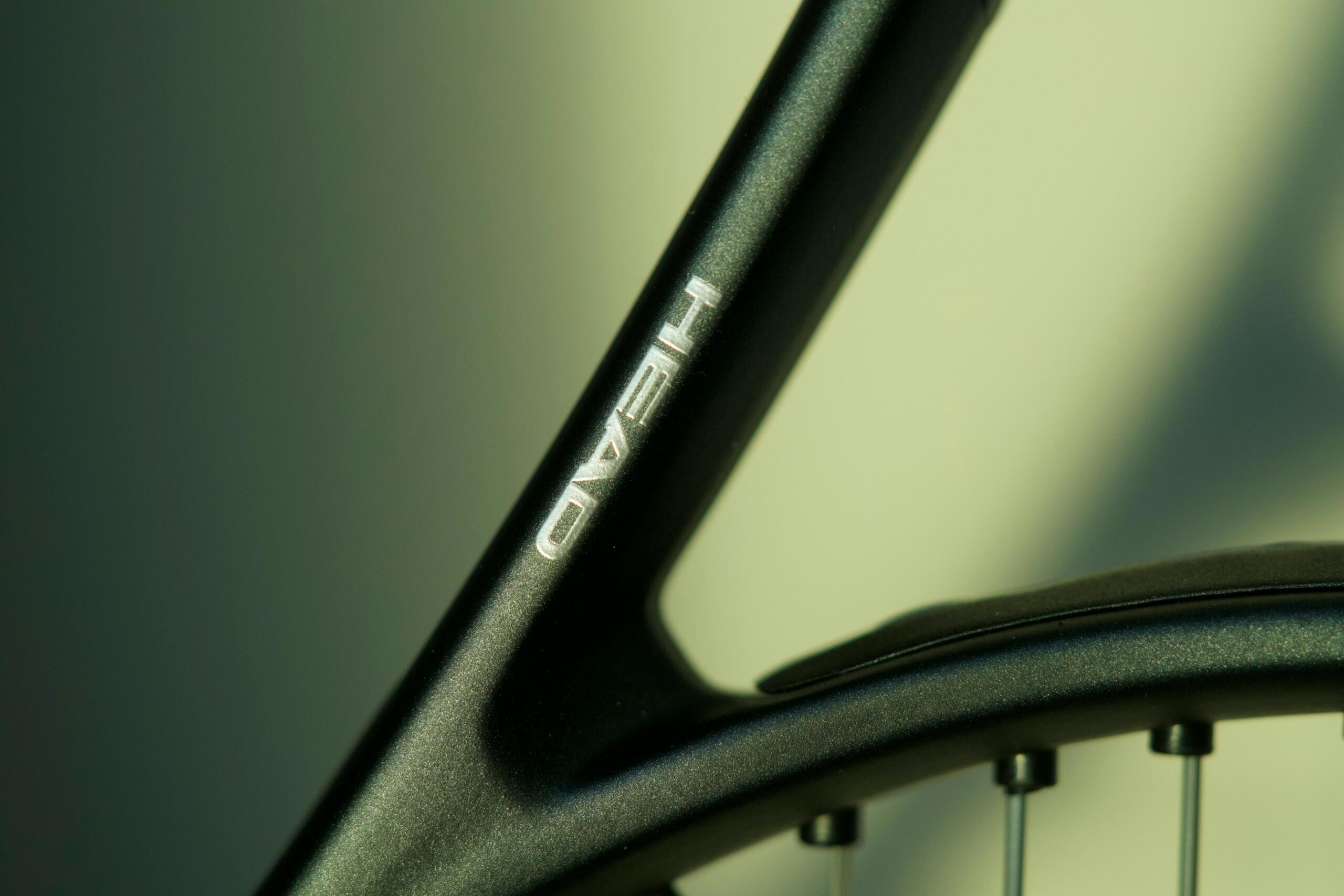 Close-up van het zwarte frame van een tennisracket met een zilverkleurig logo en een deel van de snaren.