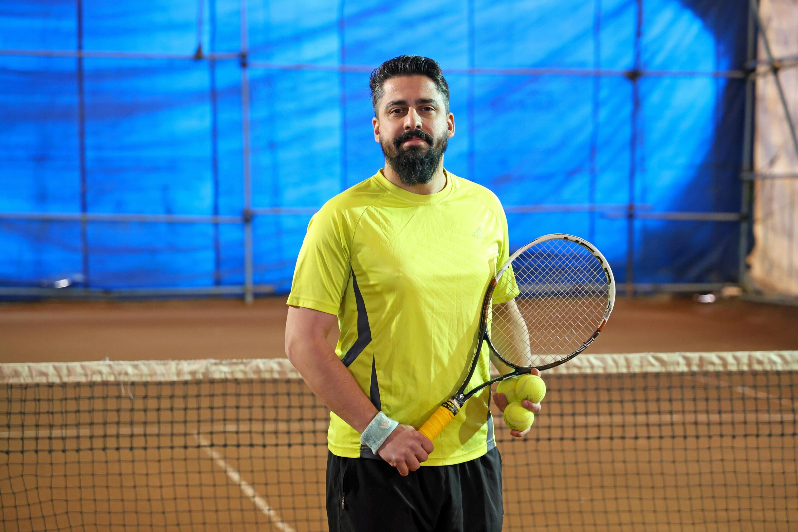 Een man in een geel shirt staat bij een tennisnet met een racket en meerdere tennisballen in zijn handen.