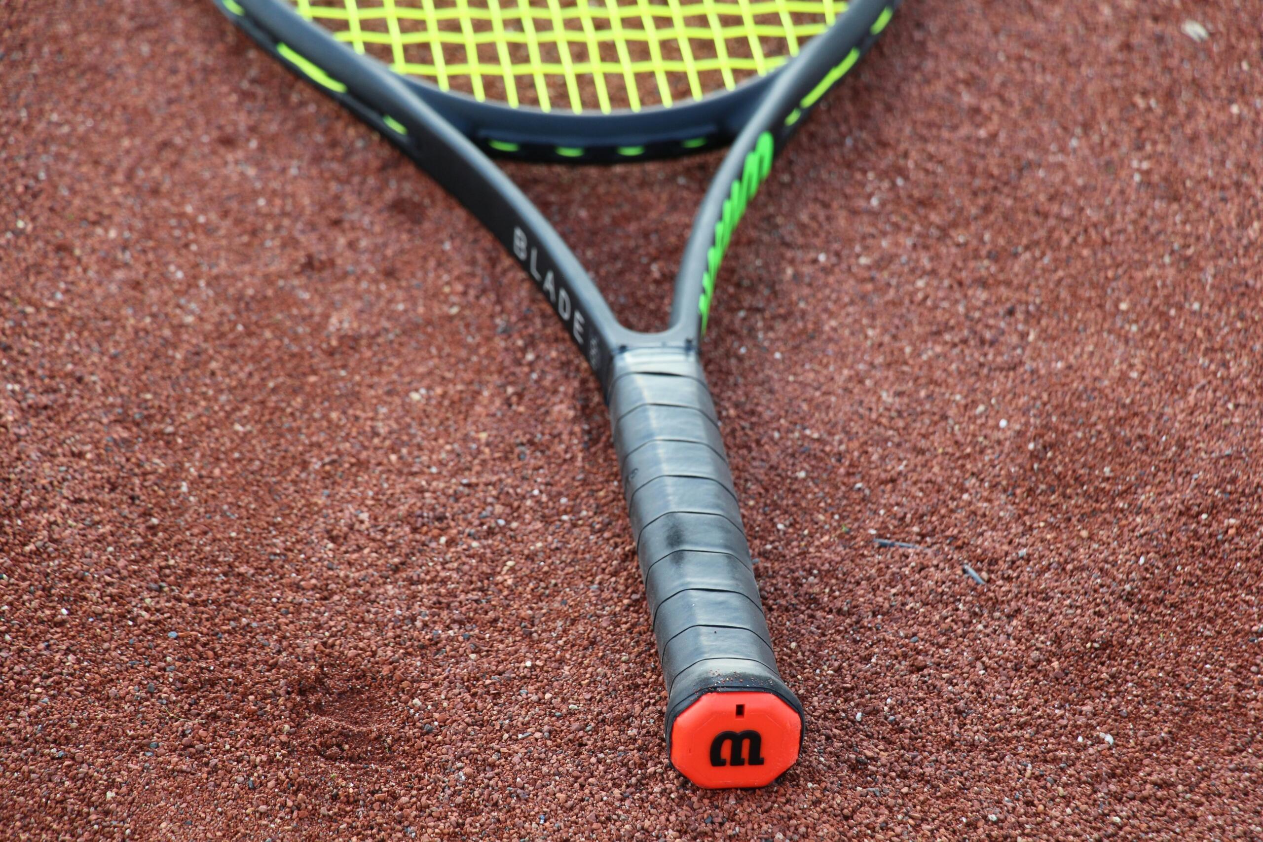 Een zwart tennisracket met groene details ligt op een gravelbaan, met het handvat naar de camera gericht.