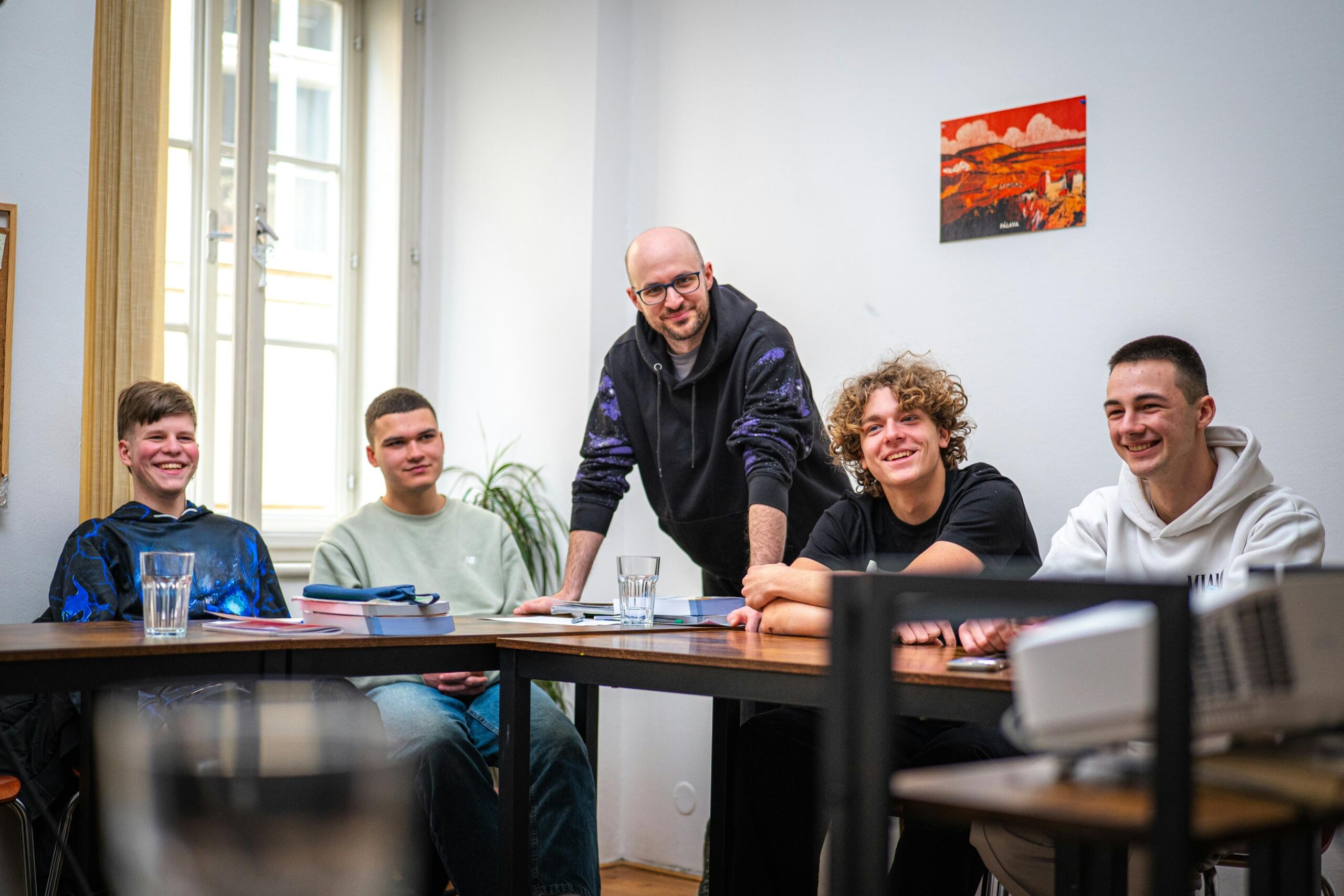 Vier jonge mannen zitten aan tafels terwijl een man achter hen staat in een lichte ruimte.