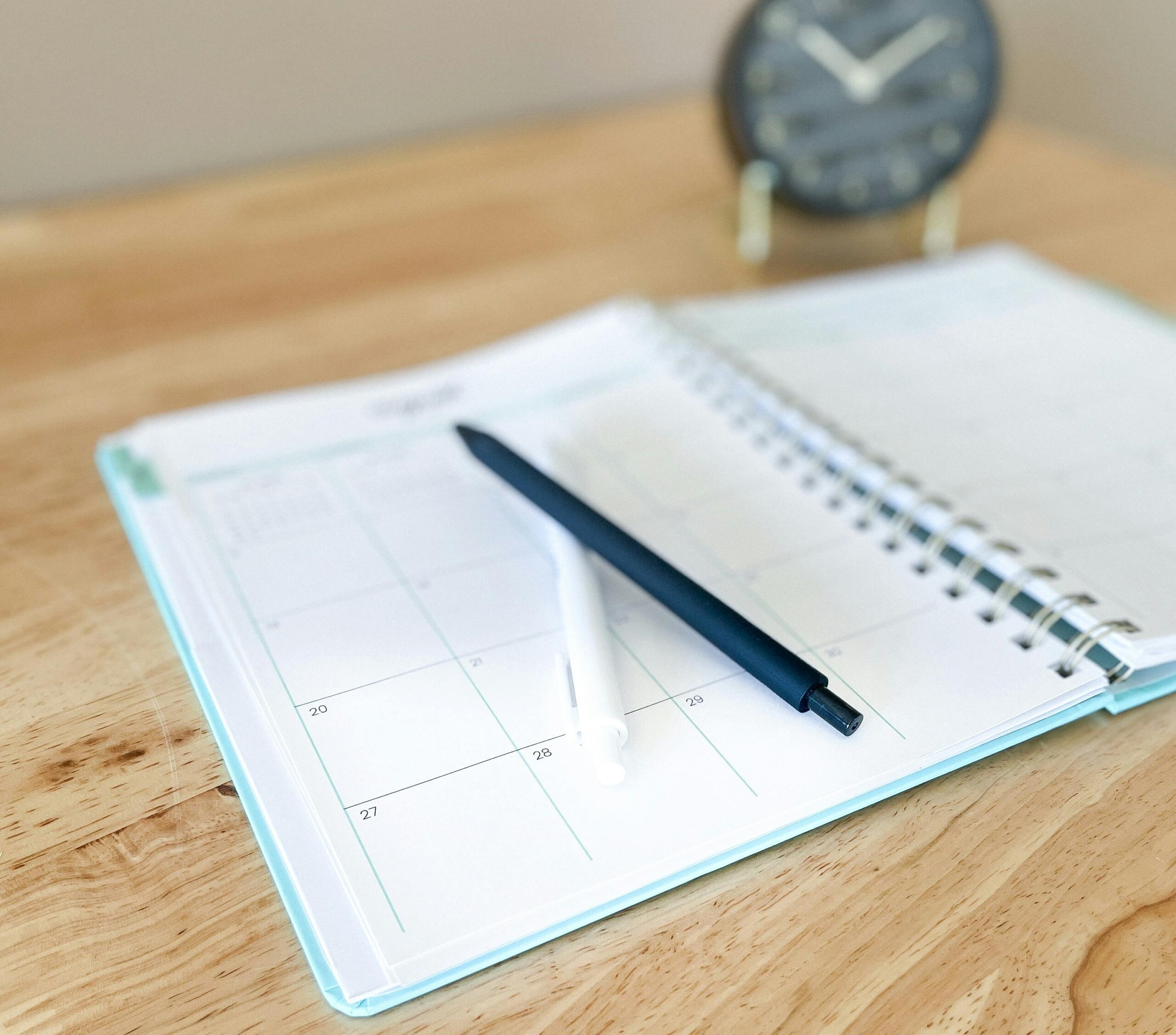 Een opengeslagen agenda met een blauwe pen ligt op een houten tafel, met een klok op de achtergrond.