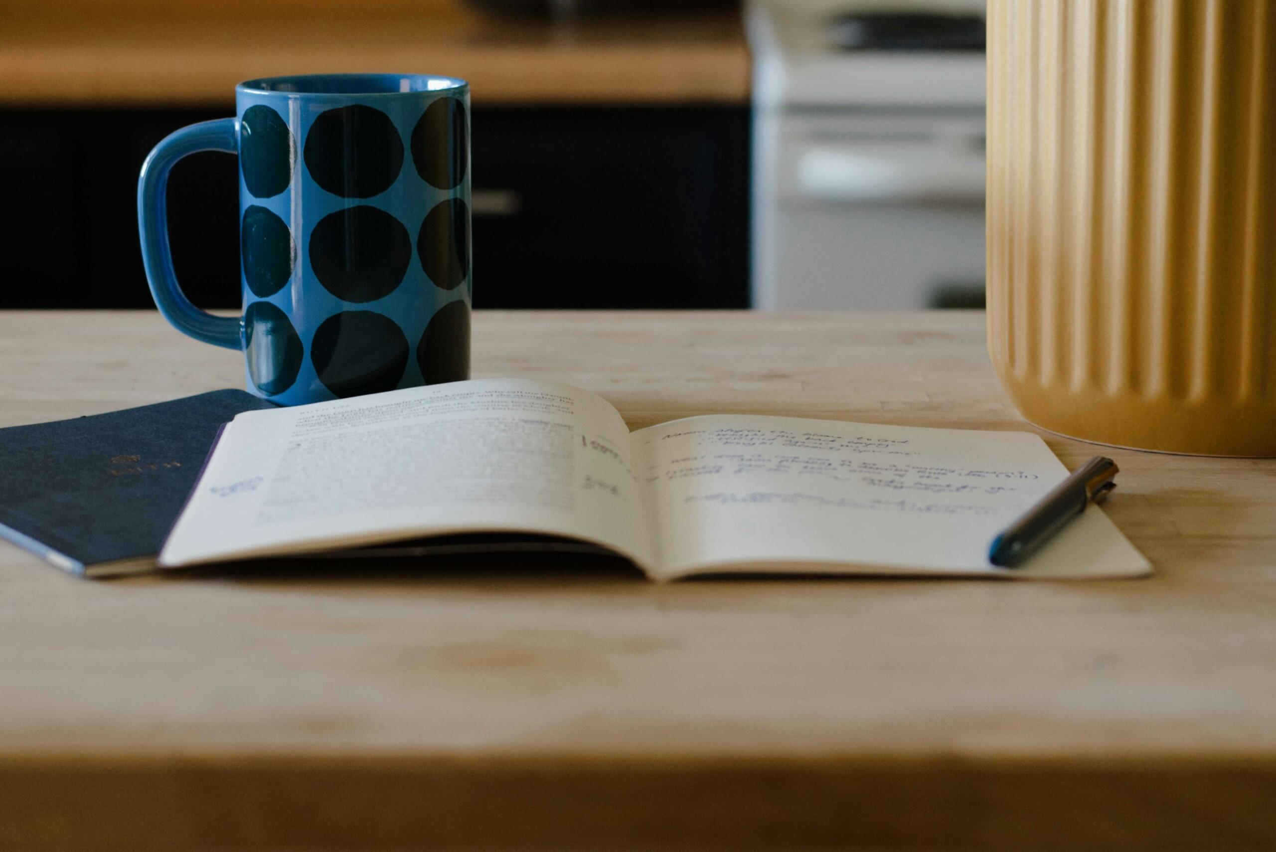 Een opengeslagen notitieboek met handgeschreven tekst ligt naast een blauwe mok en een pen op een houten tafel.