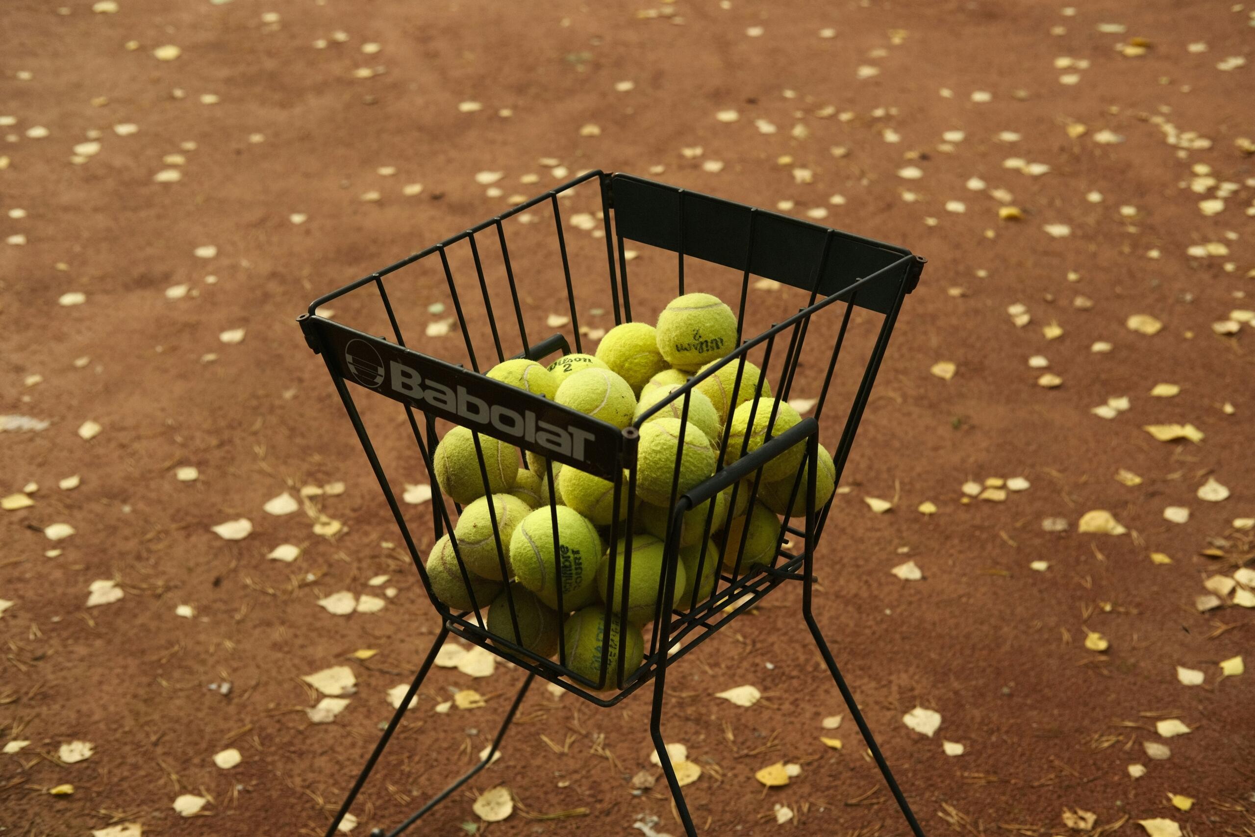 Een metalen ballenmand vol tennisballen staat op een gravelbaan tussen verspreide bladeren.