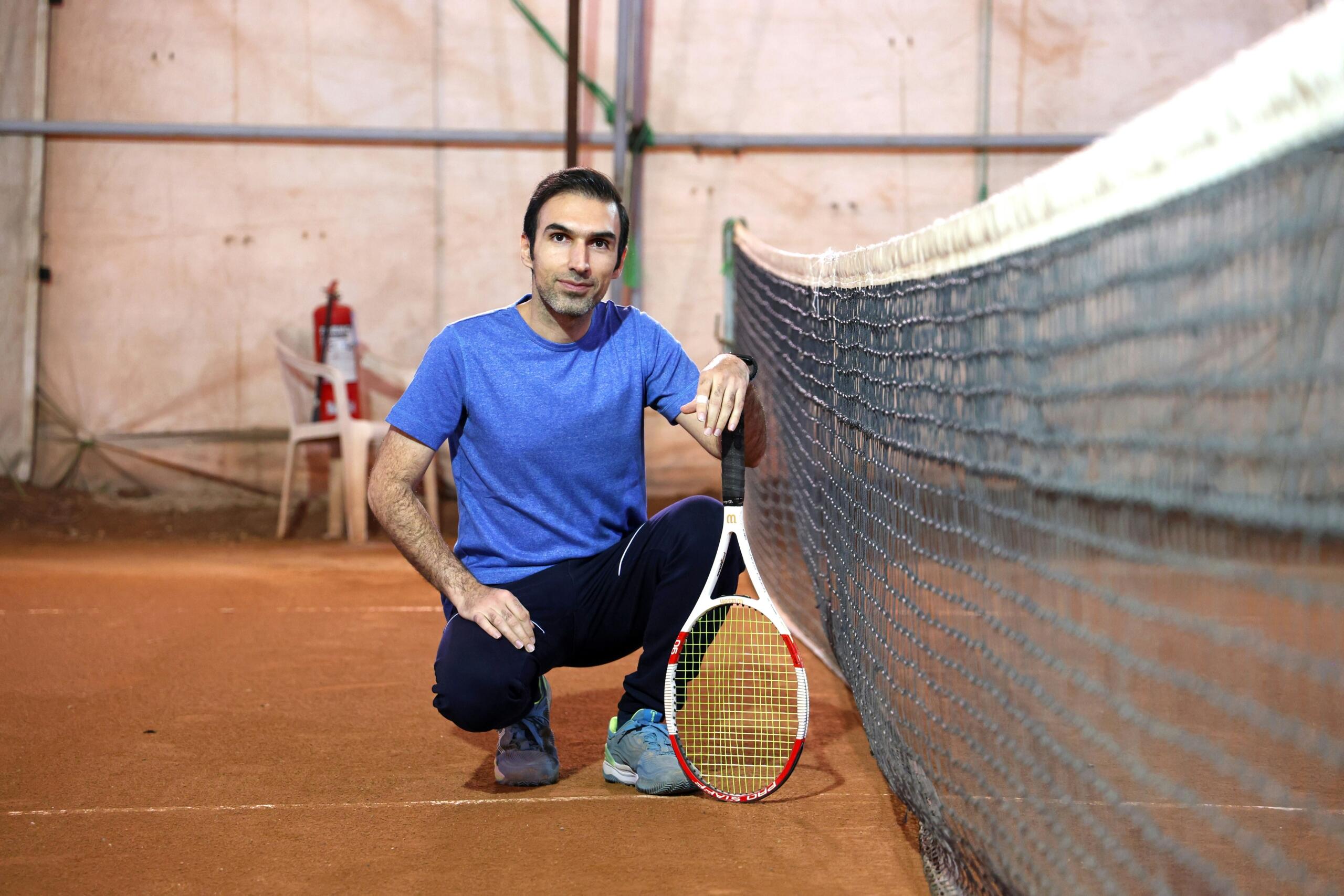 Een man knielt naast een tennisnet en houdt een racket rechtop voor zich op een gravelbaan.
