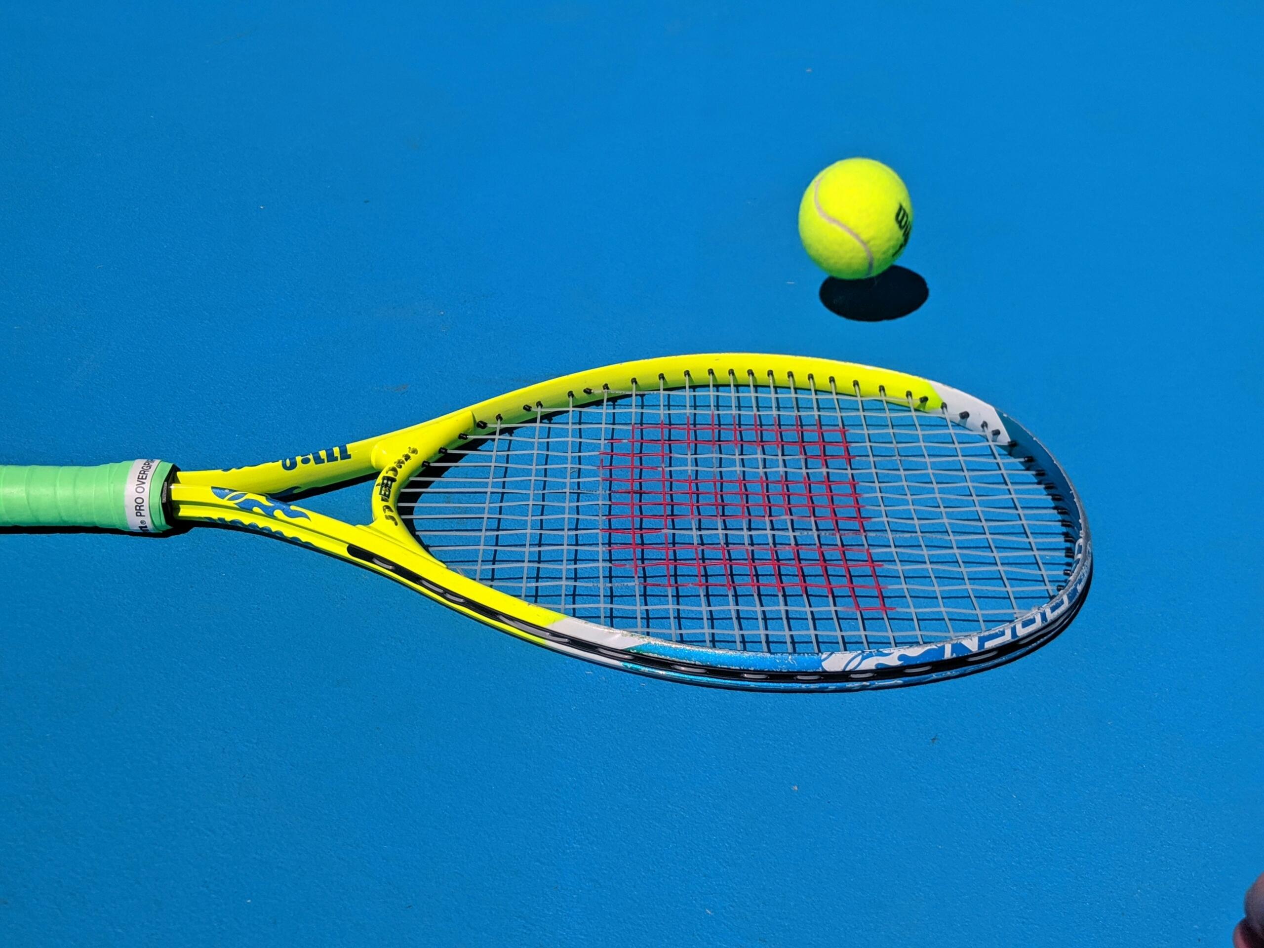 Een geel tennisracket en een tennisbal liggen op een blauwe tennisbaan.