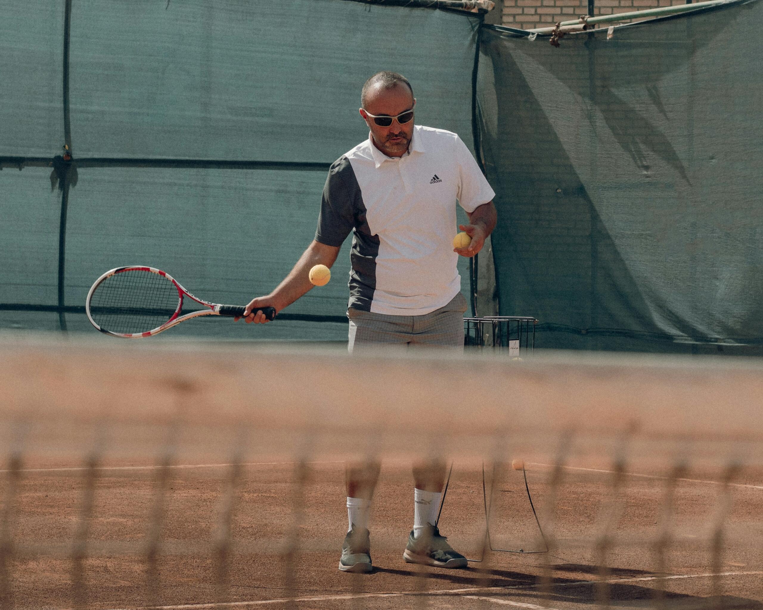 Een man met een zonnebril houdt een tennisracket en twee tennisballen vast op een gravelbaan, met een ballenmand naast zich.