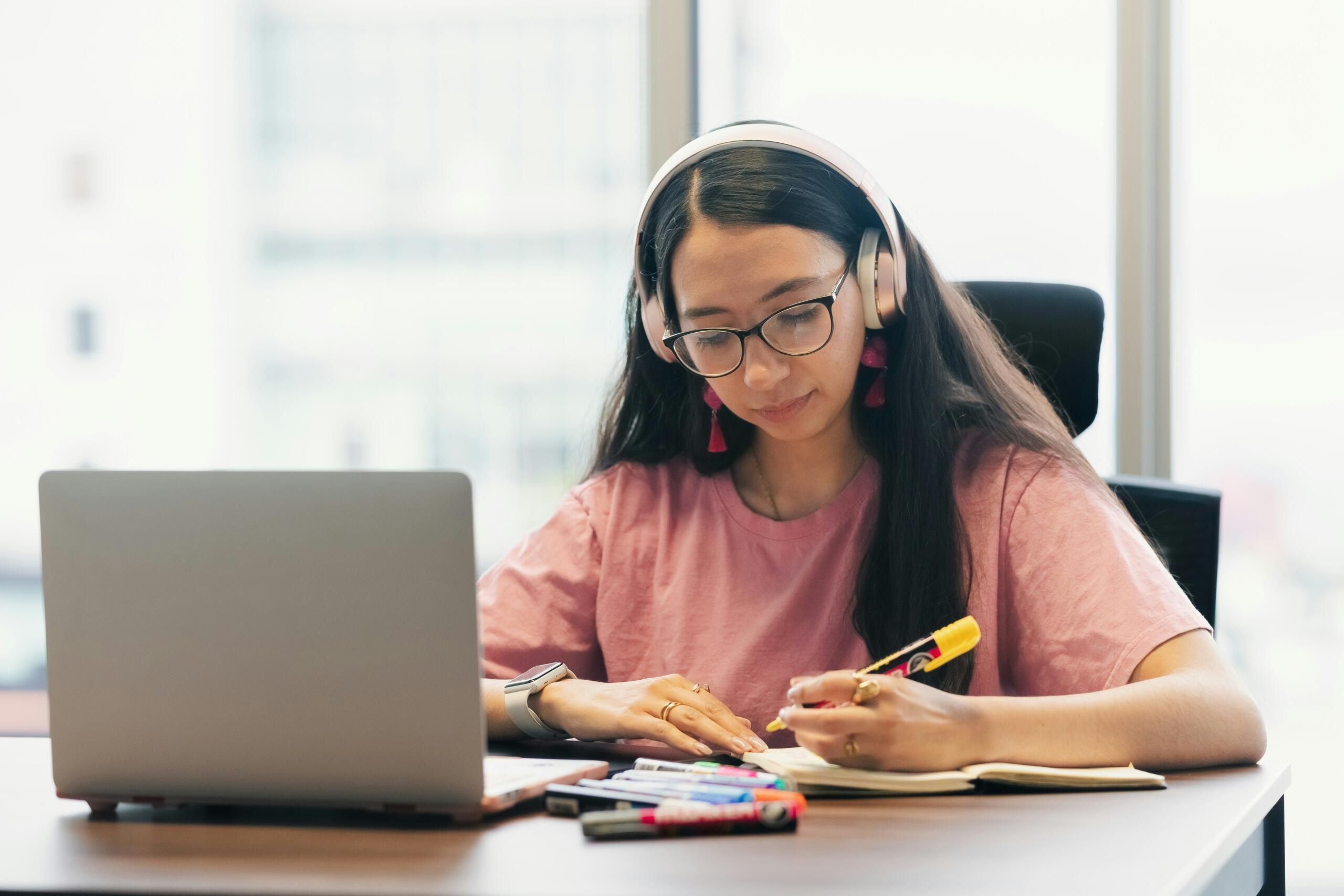 Een vrouw met een koptelefoon schrijft in een notitieboek naast een geopende laptop.