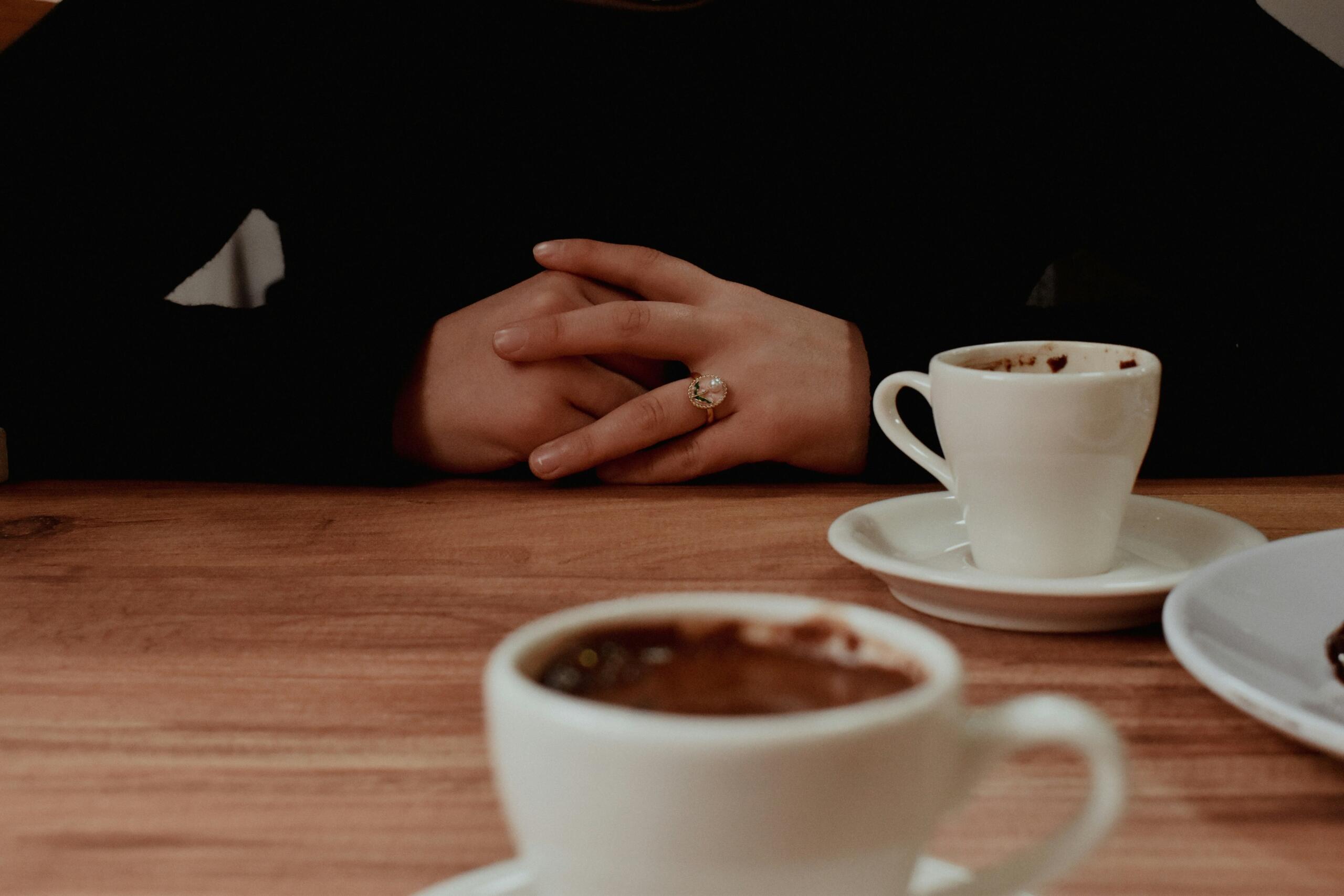 Twee kopjes koffie staan op een houten tafel, met gevouwen handen op de achtergrond.
