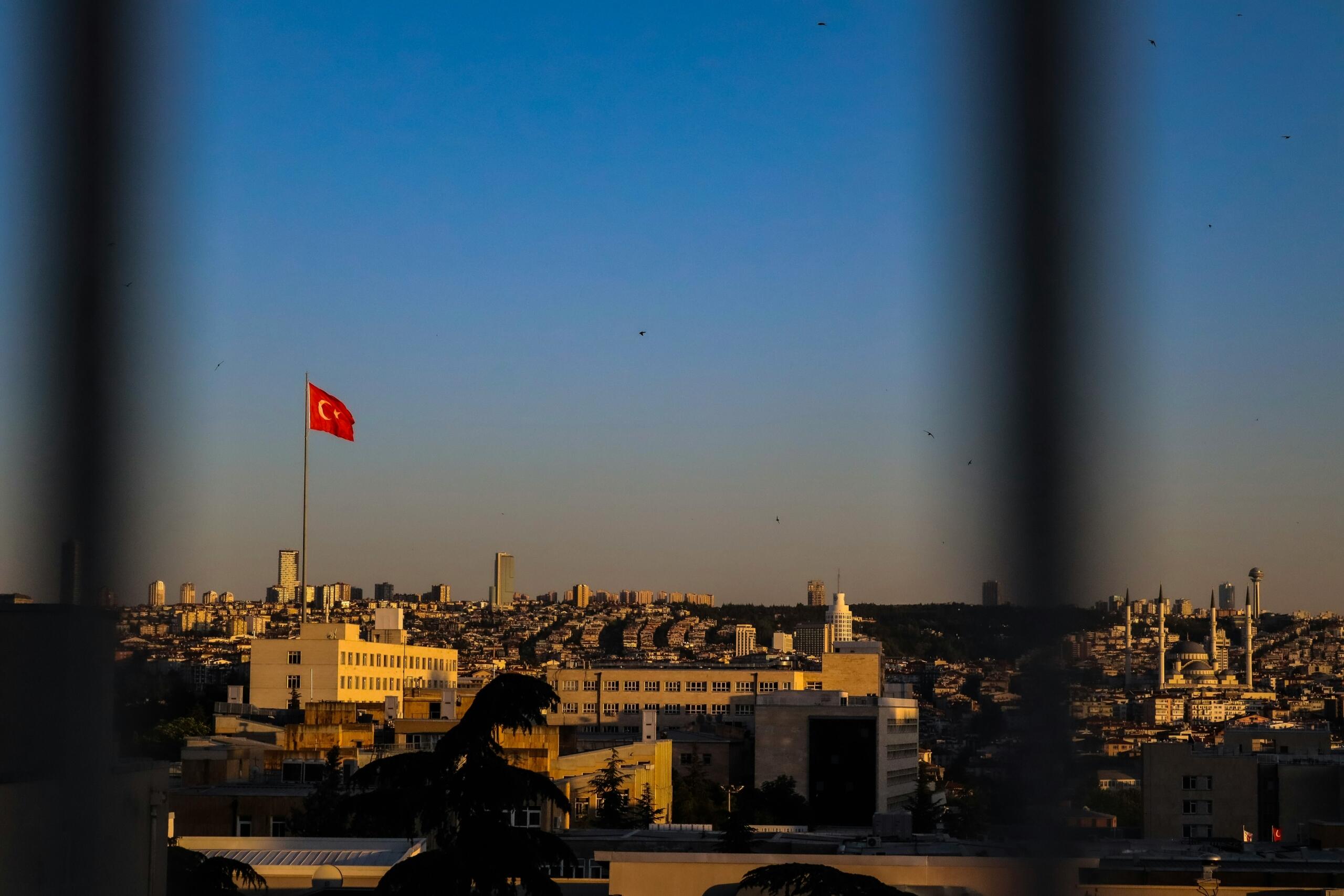 Een Turkse vlag wappert boven een stadsgezicht bij zonsondergang, gezien tussen donkere spijlen door.