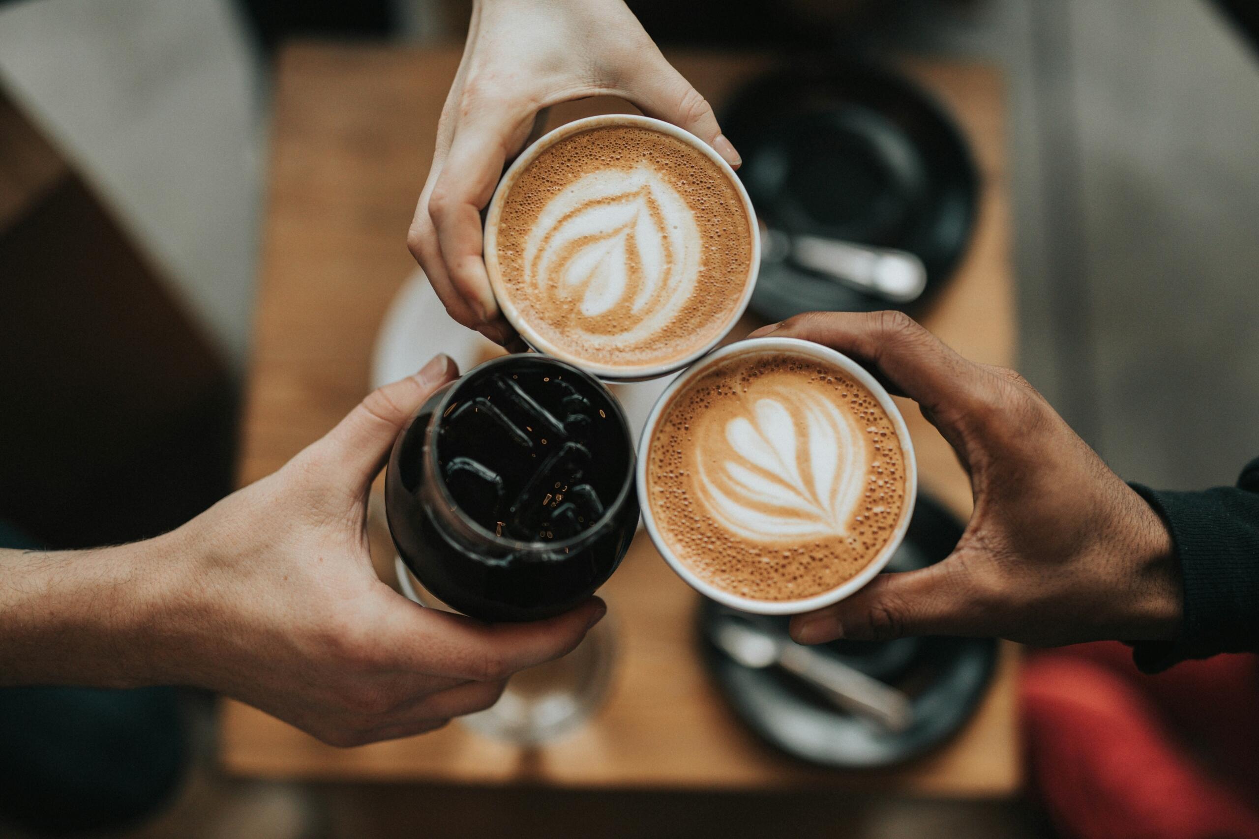 Drie handen houden twee koppen koffie en een glas met een donkere drank boven een tafel.