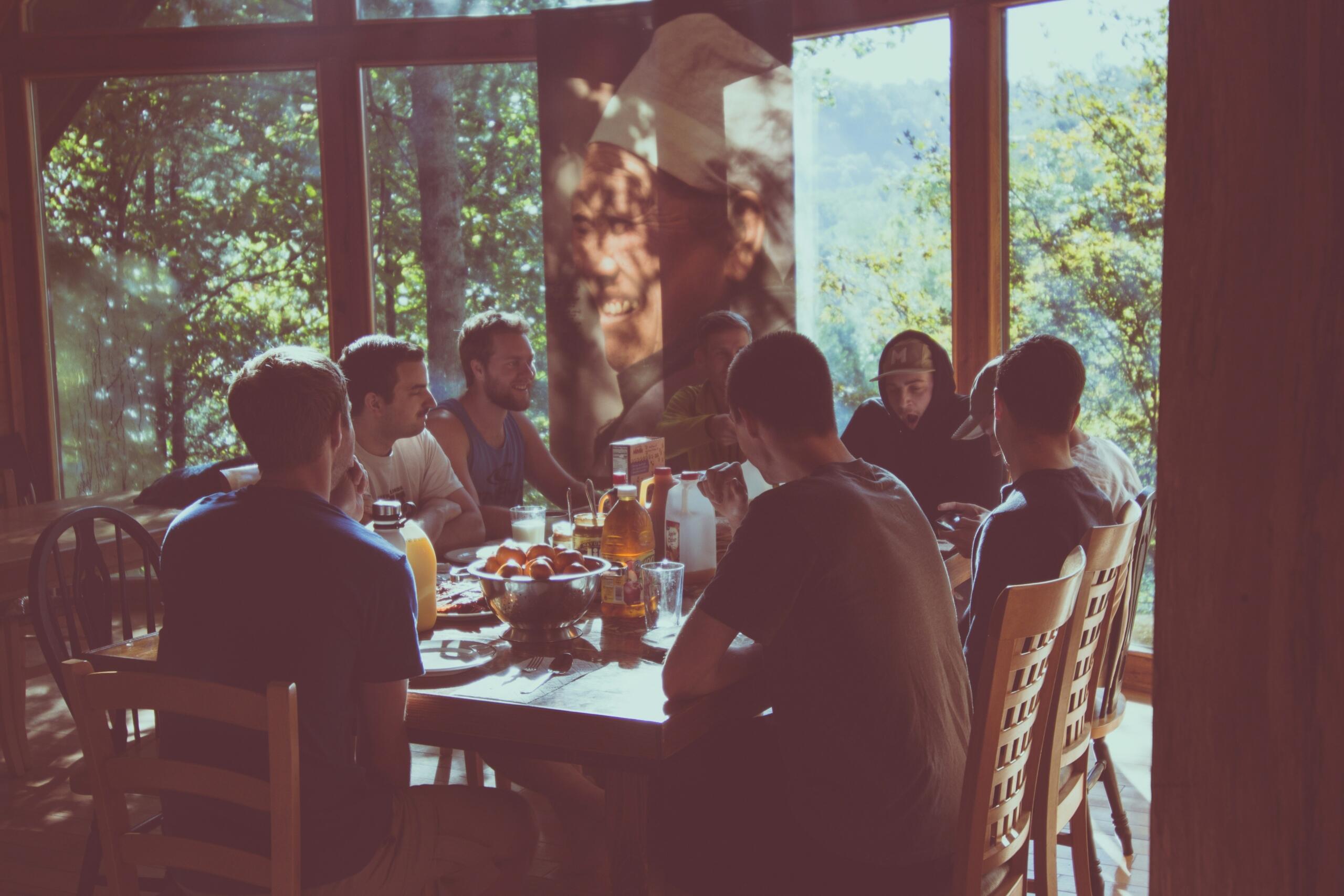 Een groep mensen zit rond een tafel met eten en drinken in een lichte ruimte.