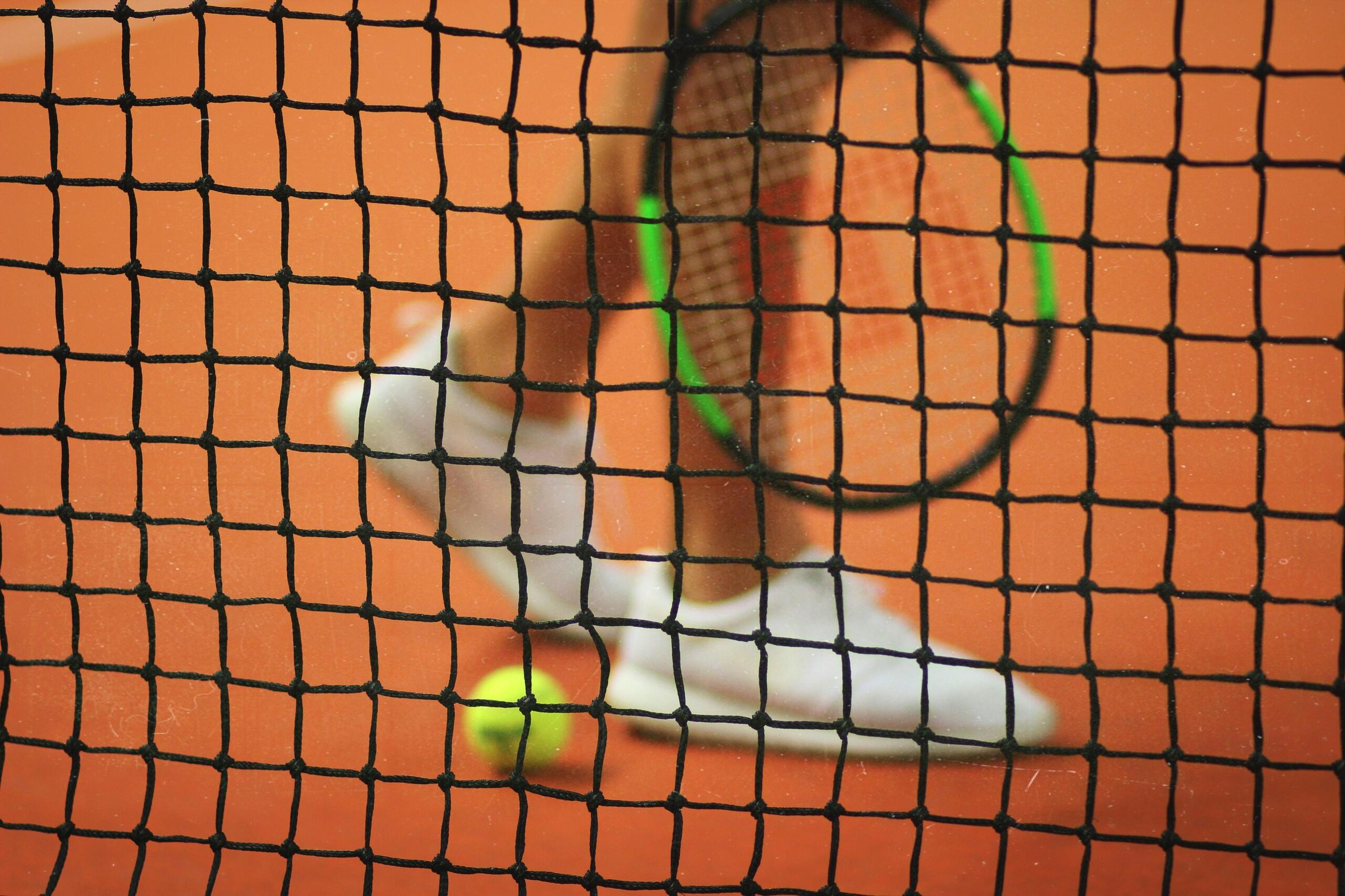 Een tennisnet staat op de voorgrond, met daarachter witte tennisschoenen, een racket en een bal op een gravelbaan.