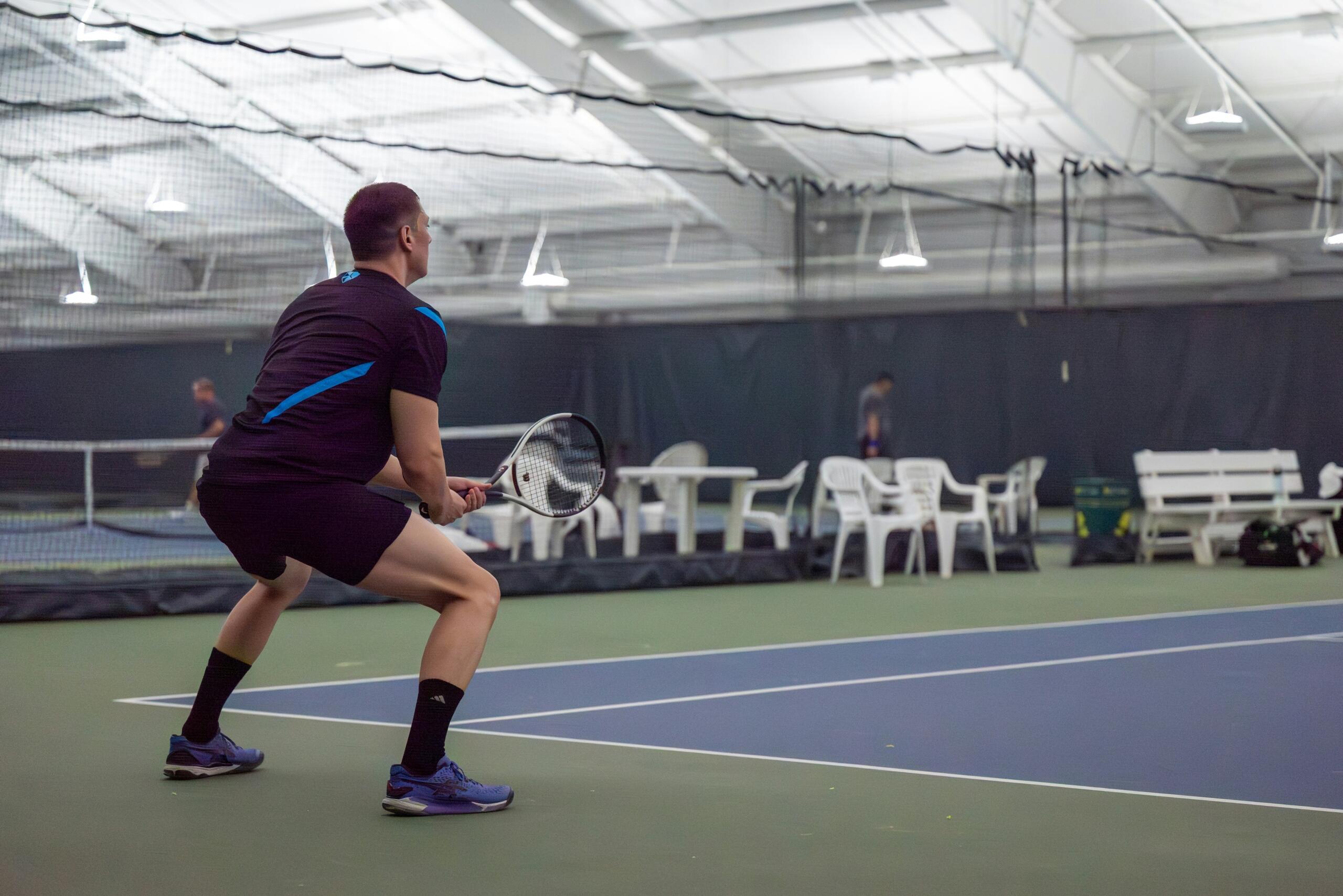 Een tennisser staat met gebogen knieën en houdt zijn racket voor zich op een overdekte tennisbaan.