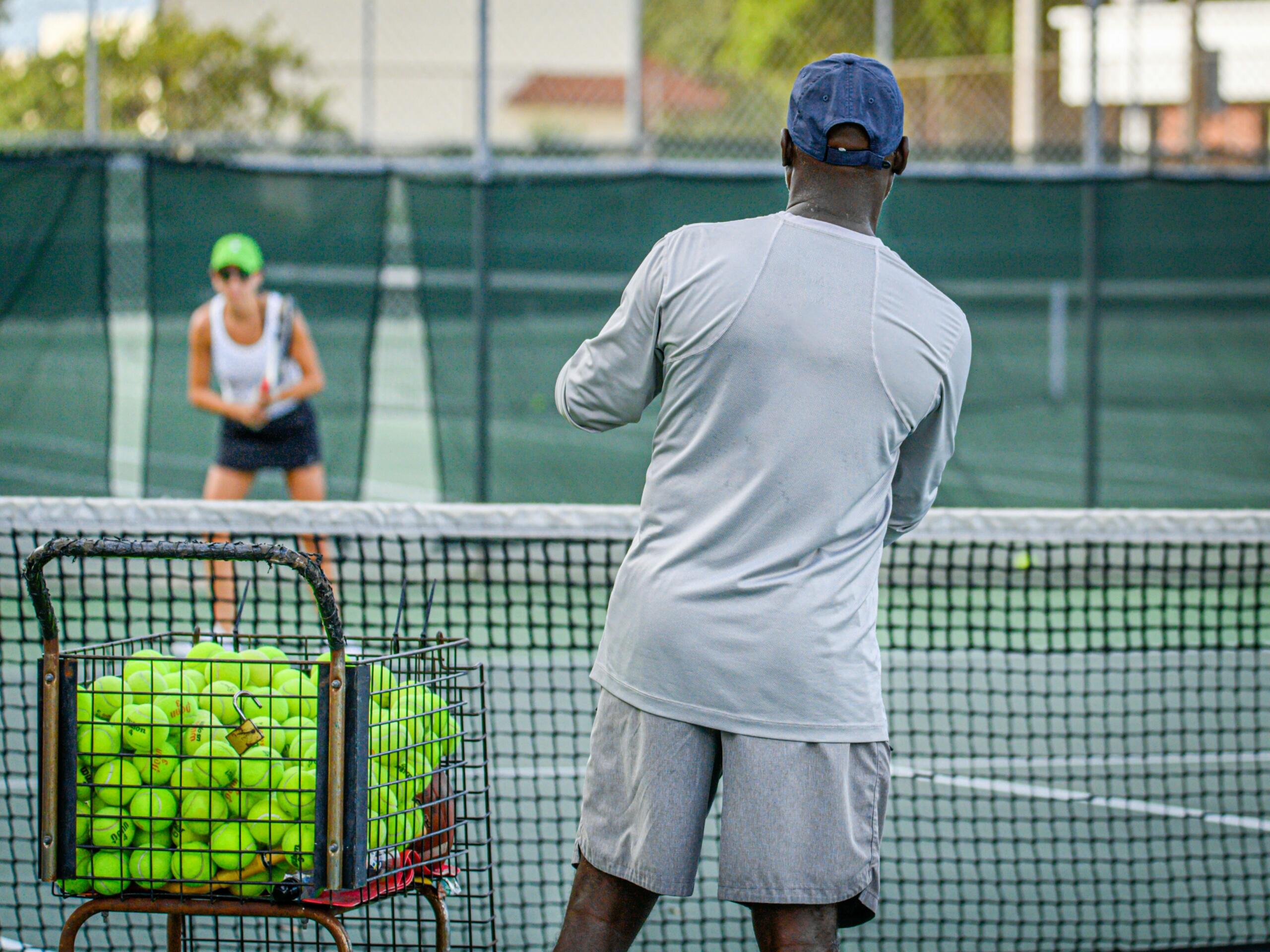 Een tenniscoach staat bij een mand met tennisballen voor het net, terwijl een speelster aan de andere kant van de baan staat.