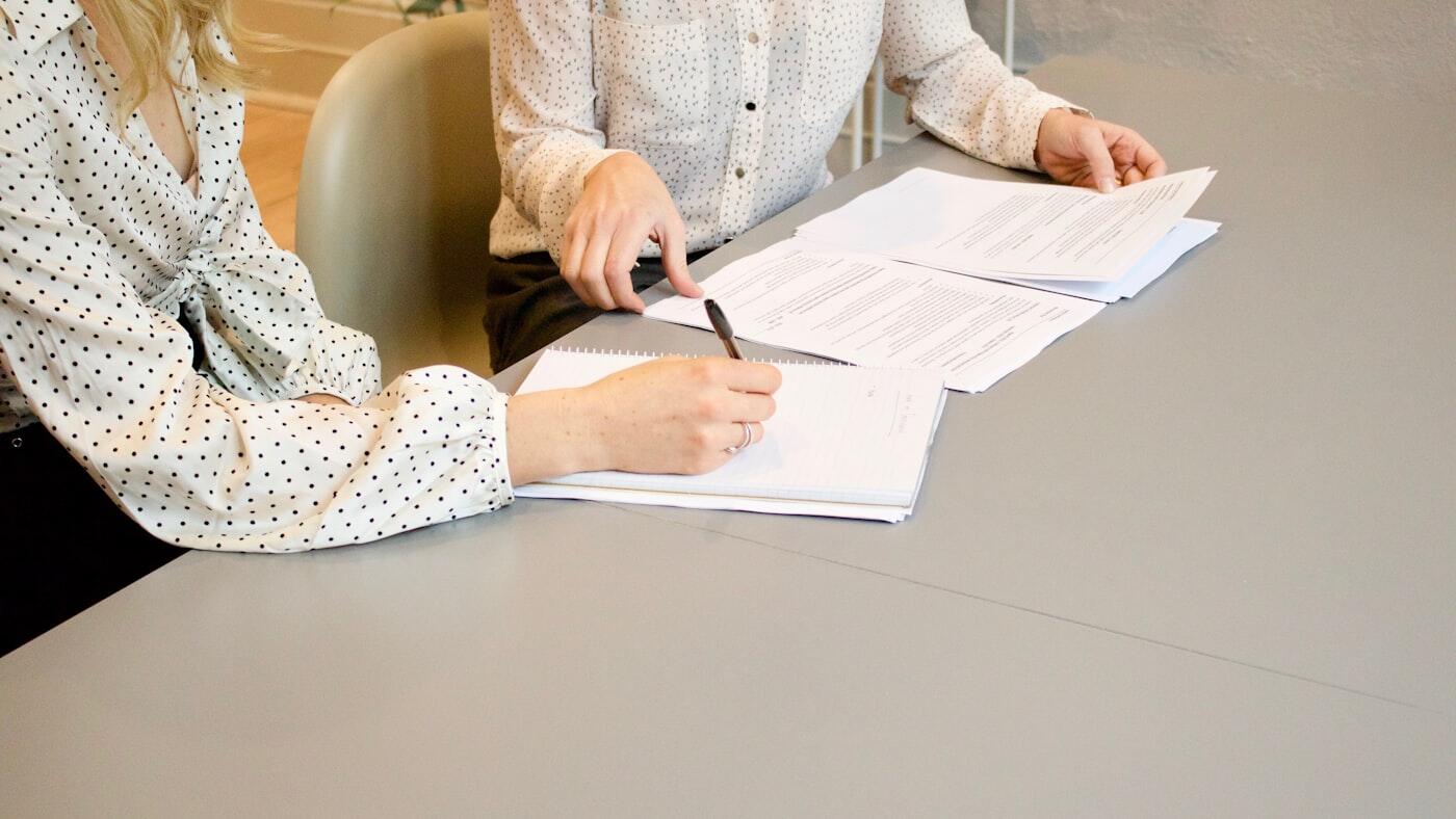 Twee personen maken aantekeningen en bekijken documenten aan een tafel.
