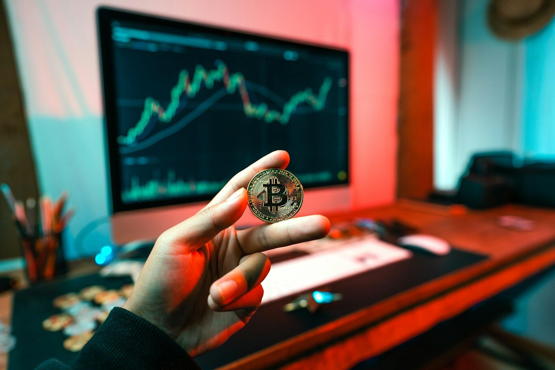 Hand houdt een bitcoinmunt voor een scherm met een koersgrafiek