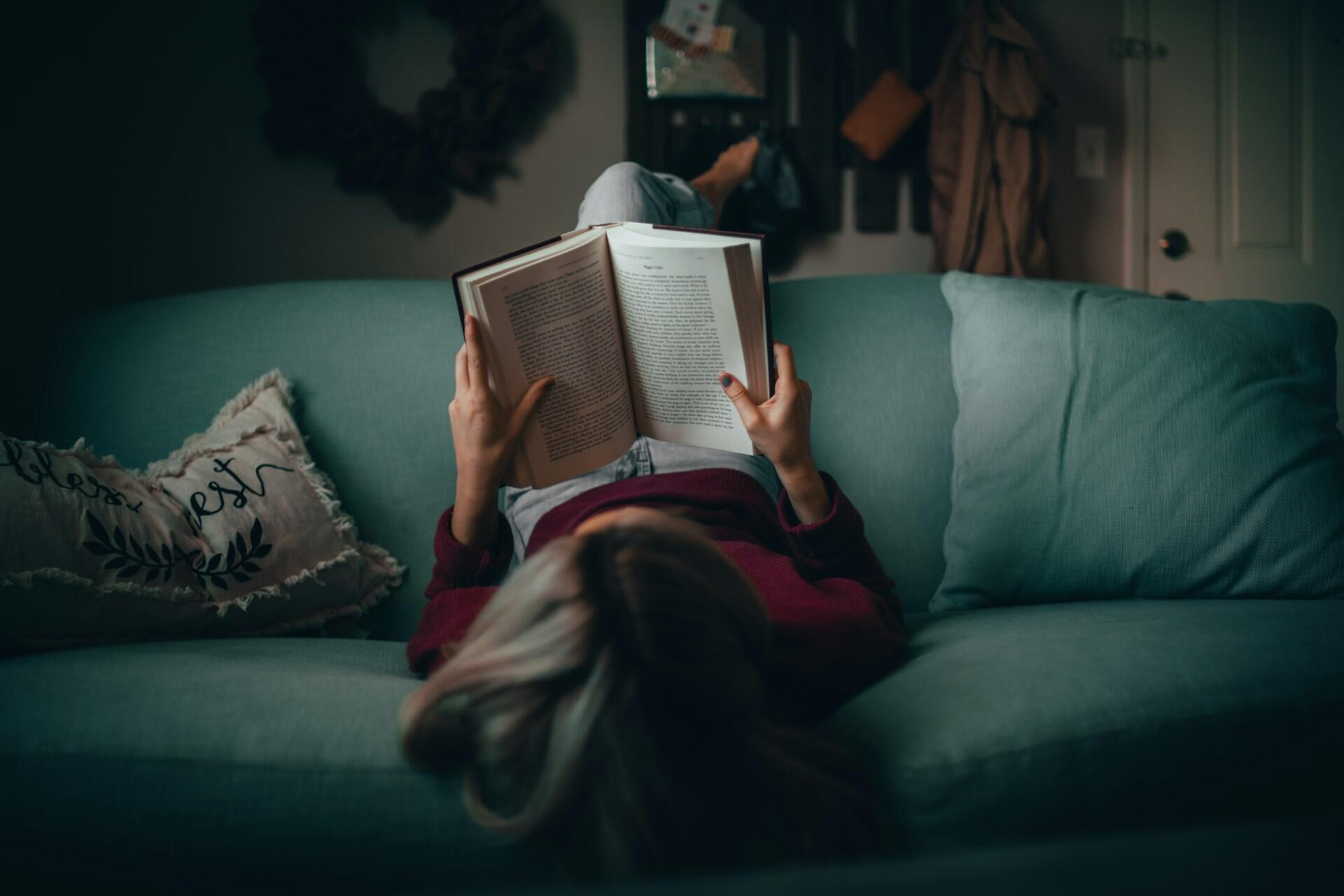 Dame ligger opp ned i grønn sofa og leser i en bok.